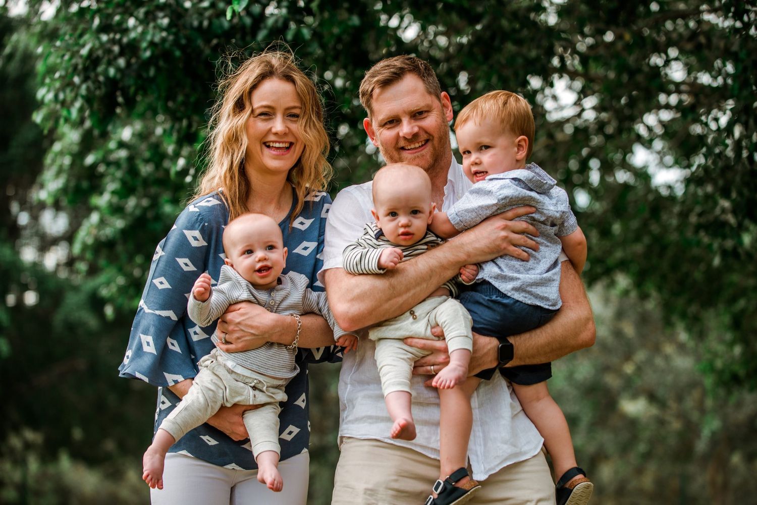 Parents holding twin babies outdoors with trees in background during family photoshoot.