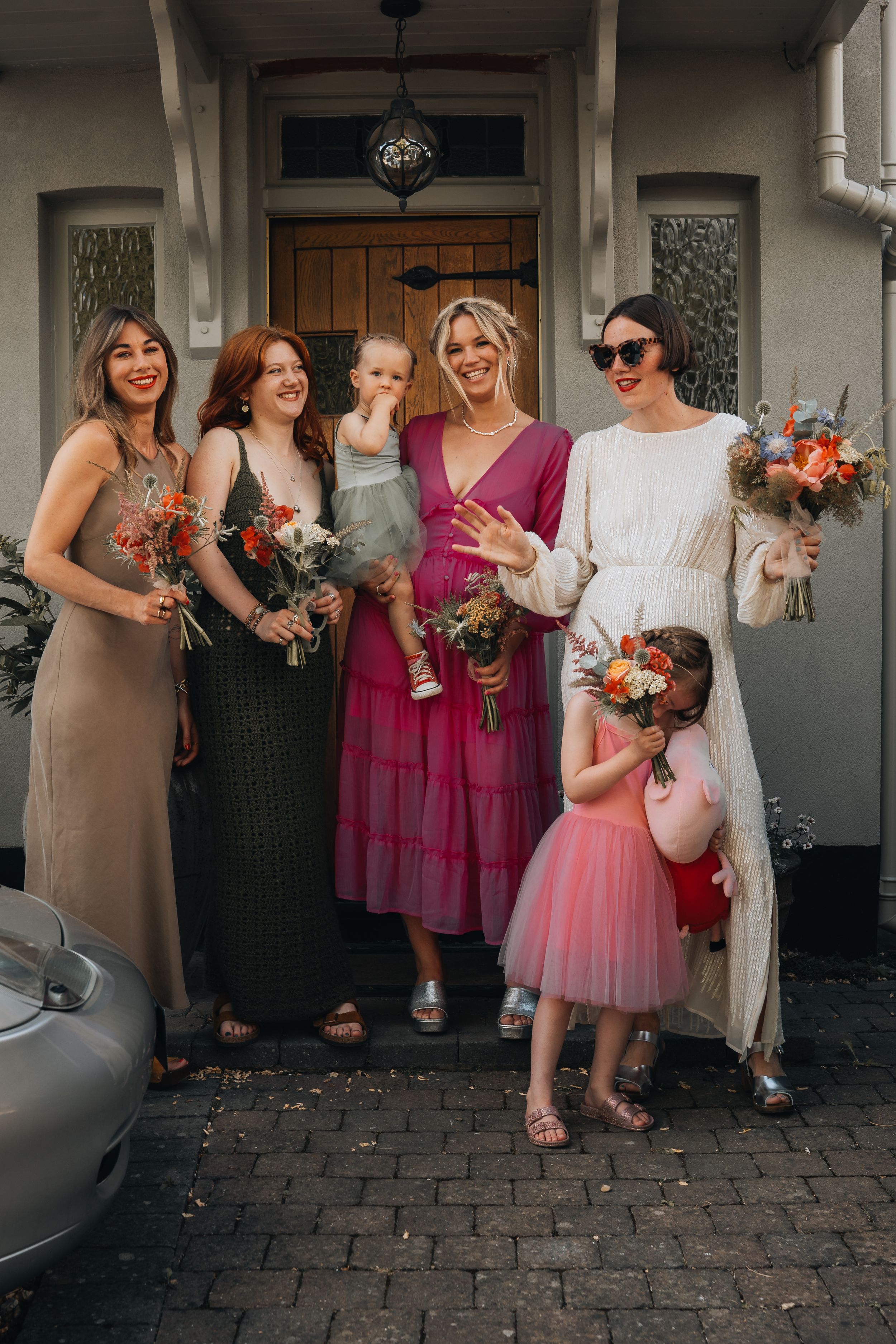 Bridesmaids in pink and neutral dresses pose together holding colorful bouquets on a cobblestone walkway.