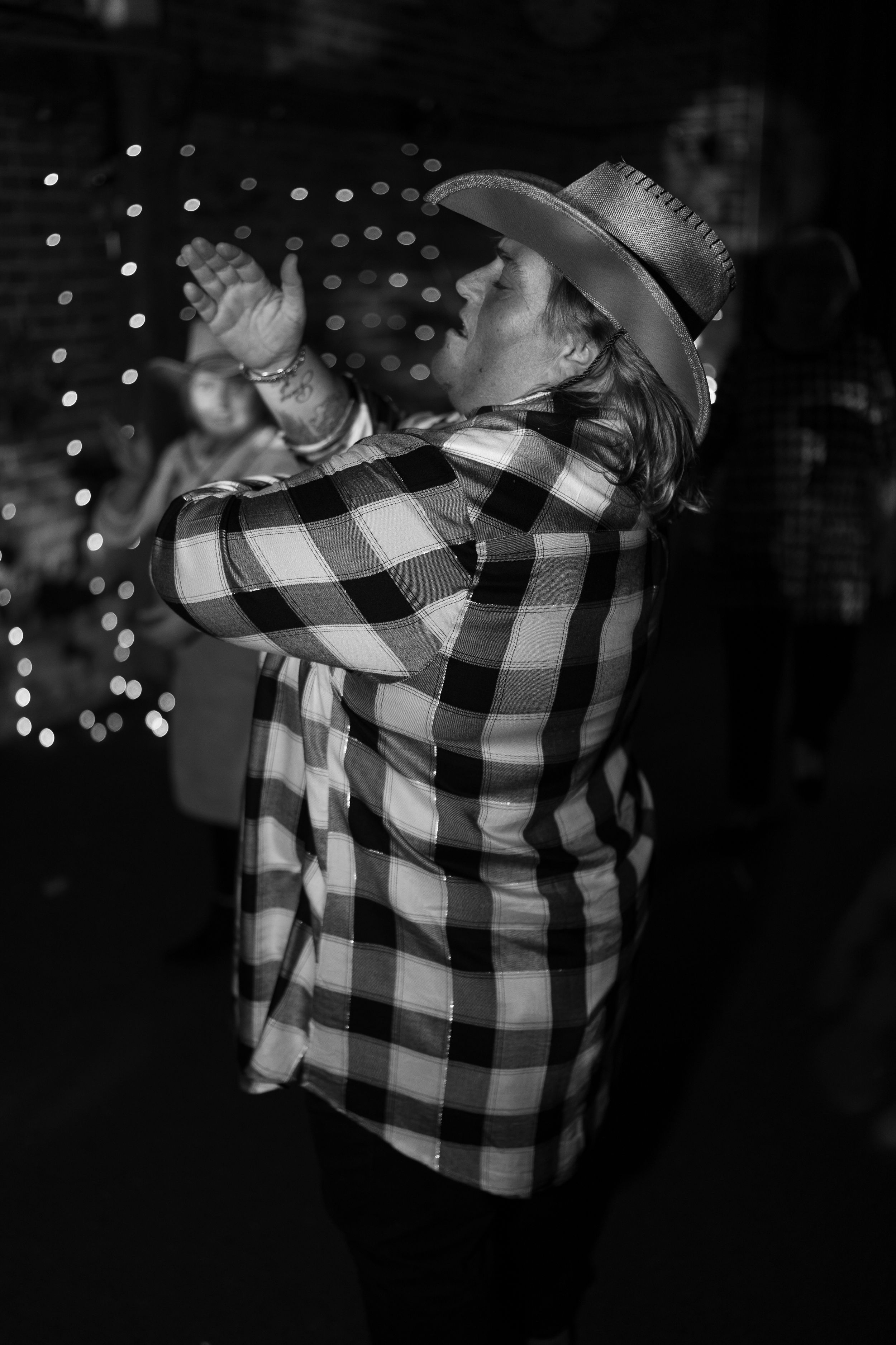 Person in black and white plaid shirt dancing in dark room with bokeh lights.