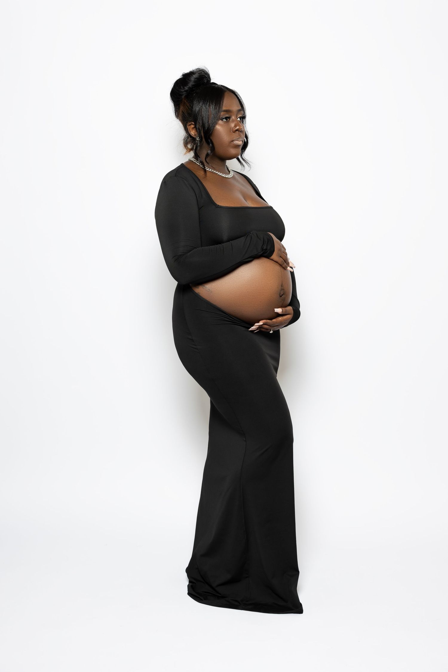 Pregnant woman in black dress poses elegantly against white background for maternity photo.