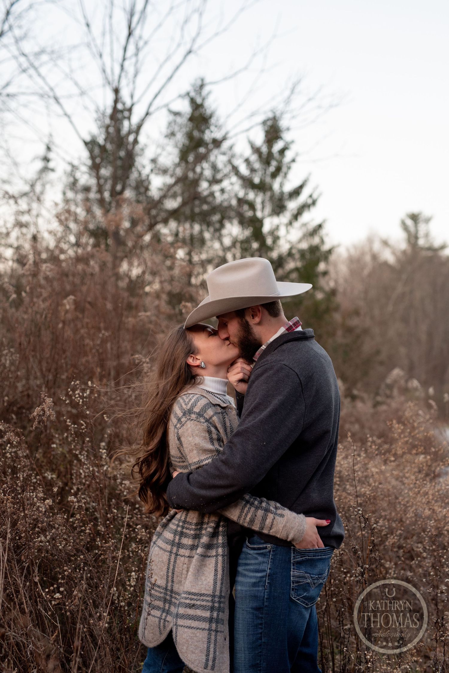 An intimate embrace between a couple wearing western attire against evergreen trees.