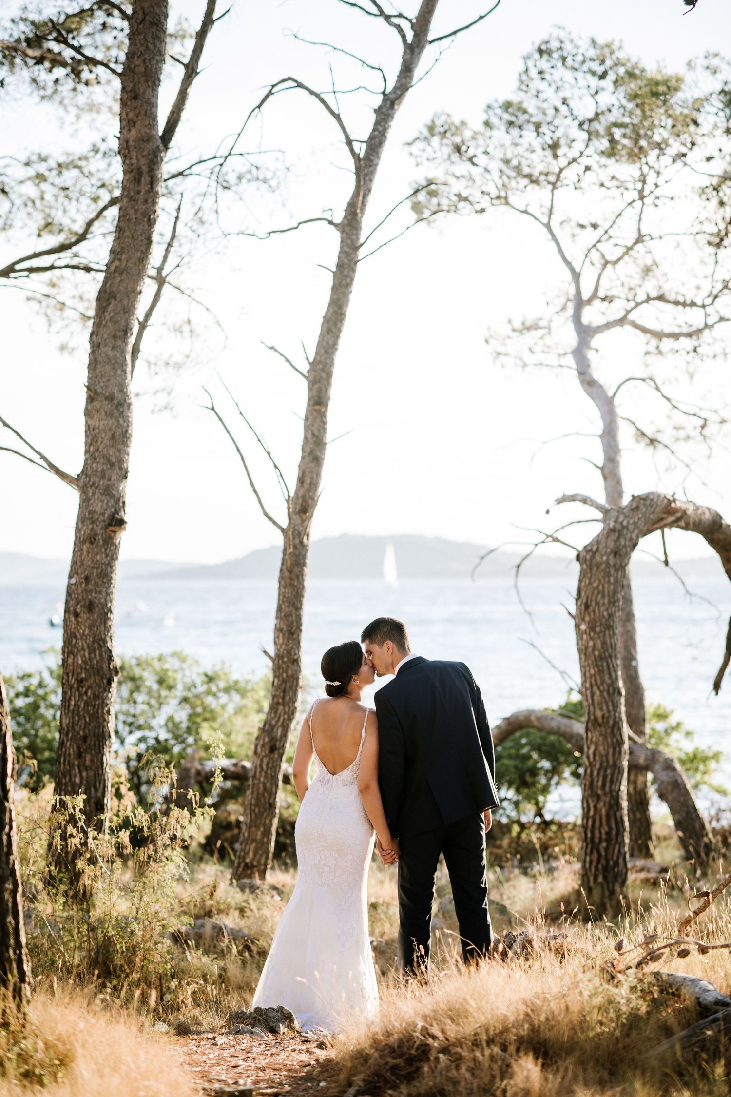 A couple in wedding attire share an intimate moment among tall trees with a sailboat visible on the water in the distance.