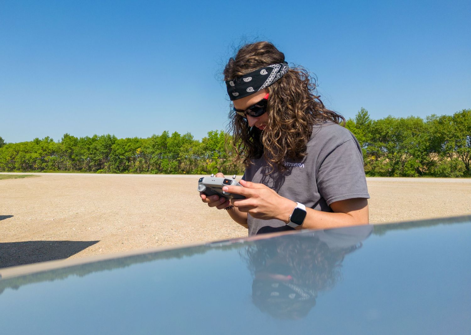 Person with long curly hair examining a camera on the hood of a car.