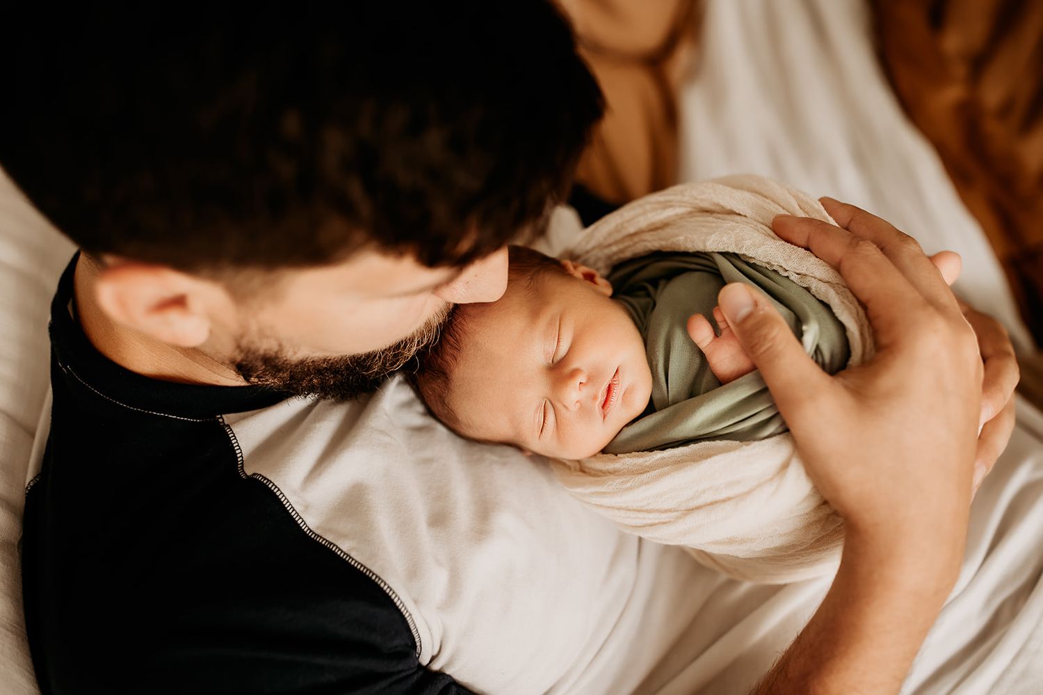 A father reclines on the bed while kissing his newborn son's head.