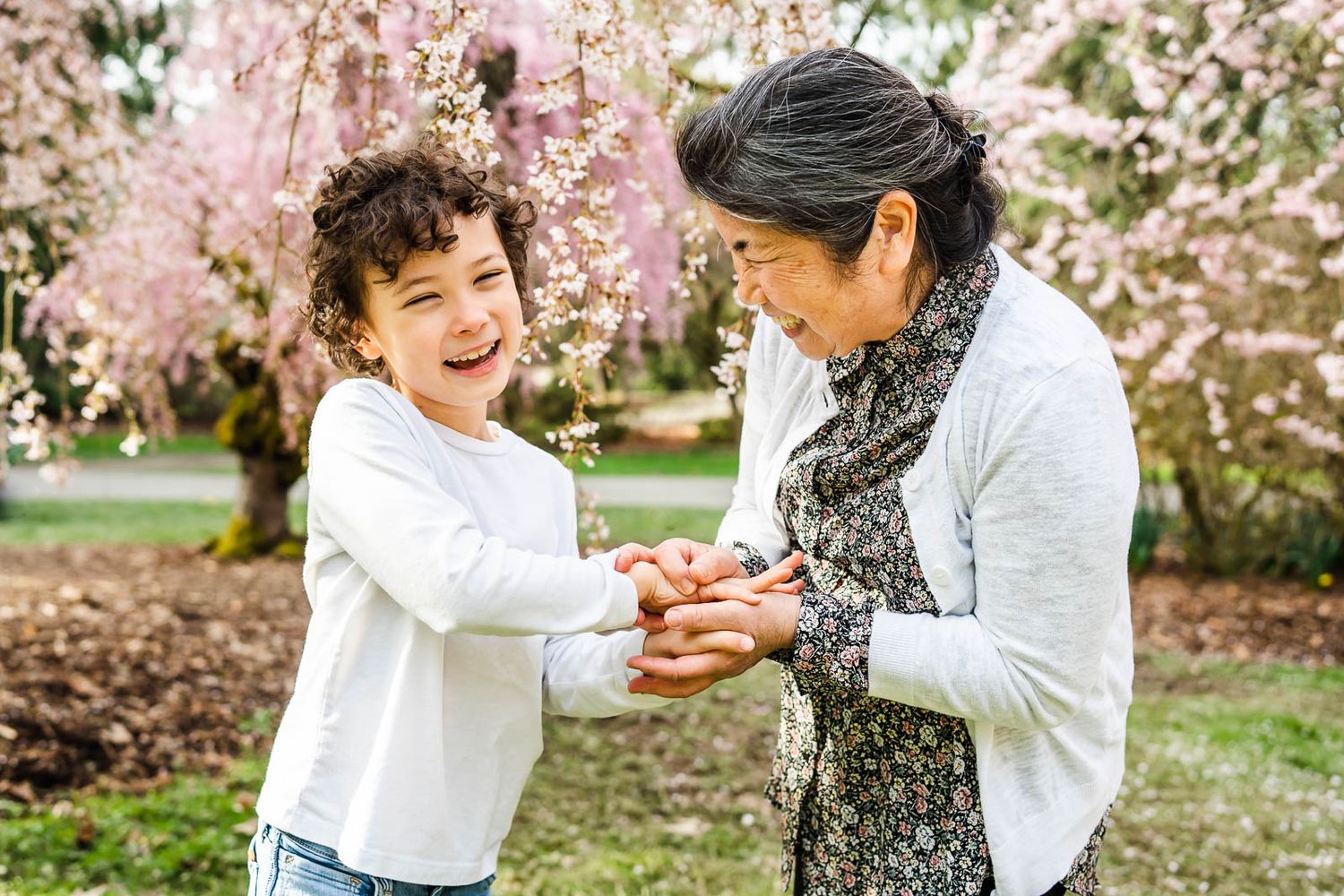 Grandmother smiling and holding grandson's hand in front of white cherry blossoms