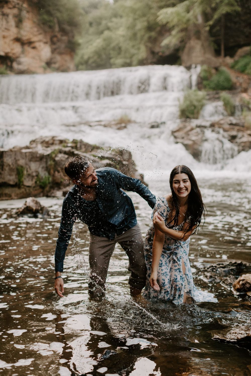 Playful couple splashes in river water during engagement photoshoot at scenic waterfall.