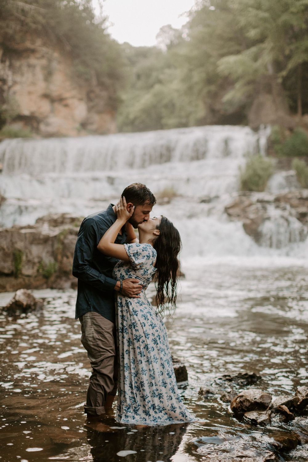 Couple embraces while standing in shallow water with cascading waterfall in background.
