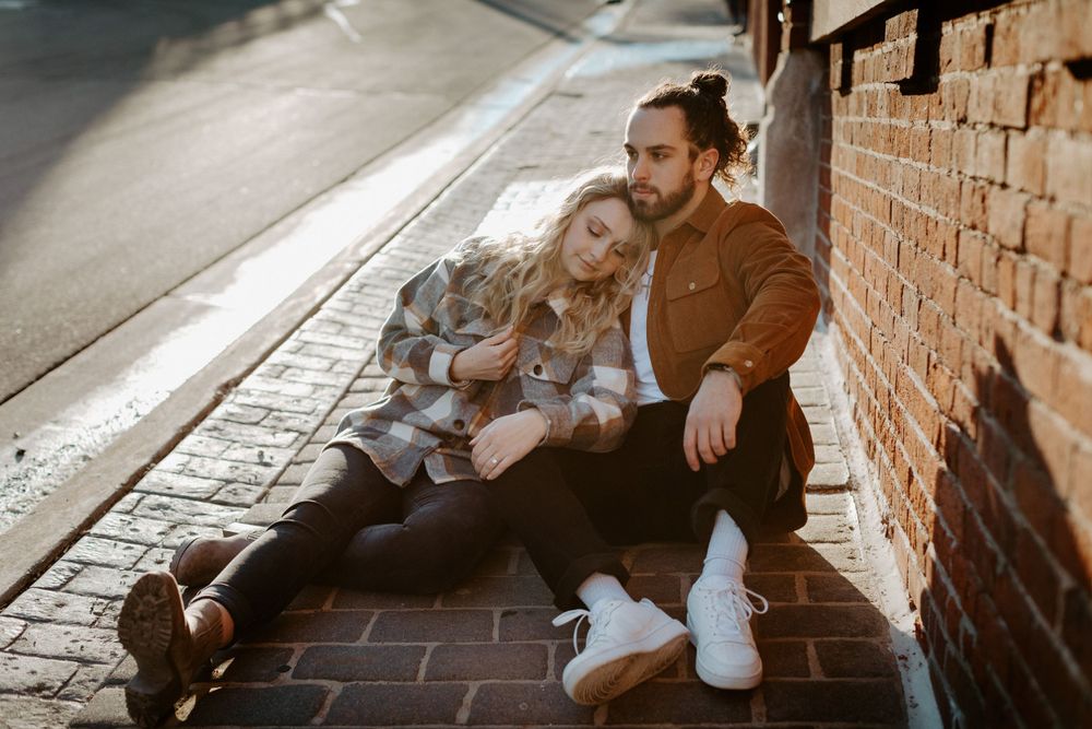 Casual urban portrait of couple sitting against brick wall on city sidewalk.
