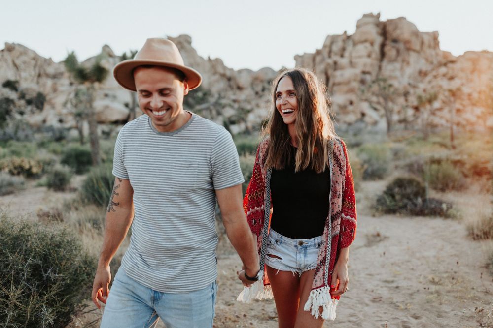 A couple laughing together on a desert hiking trail with rock formations in the background during golden hour.
