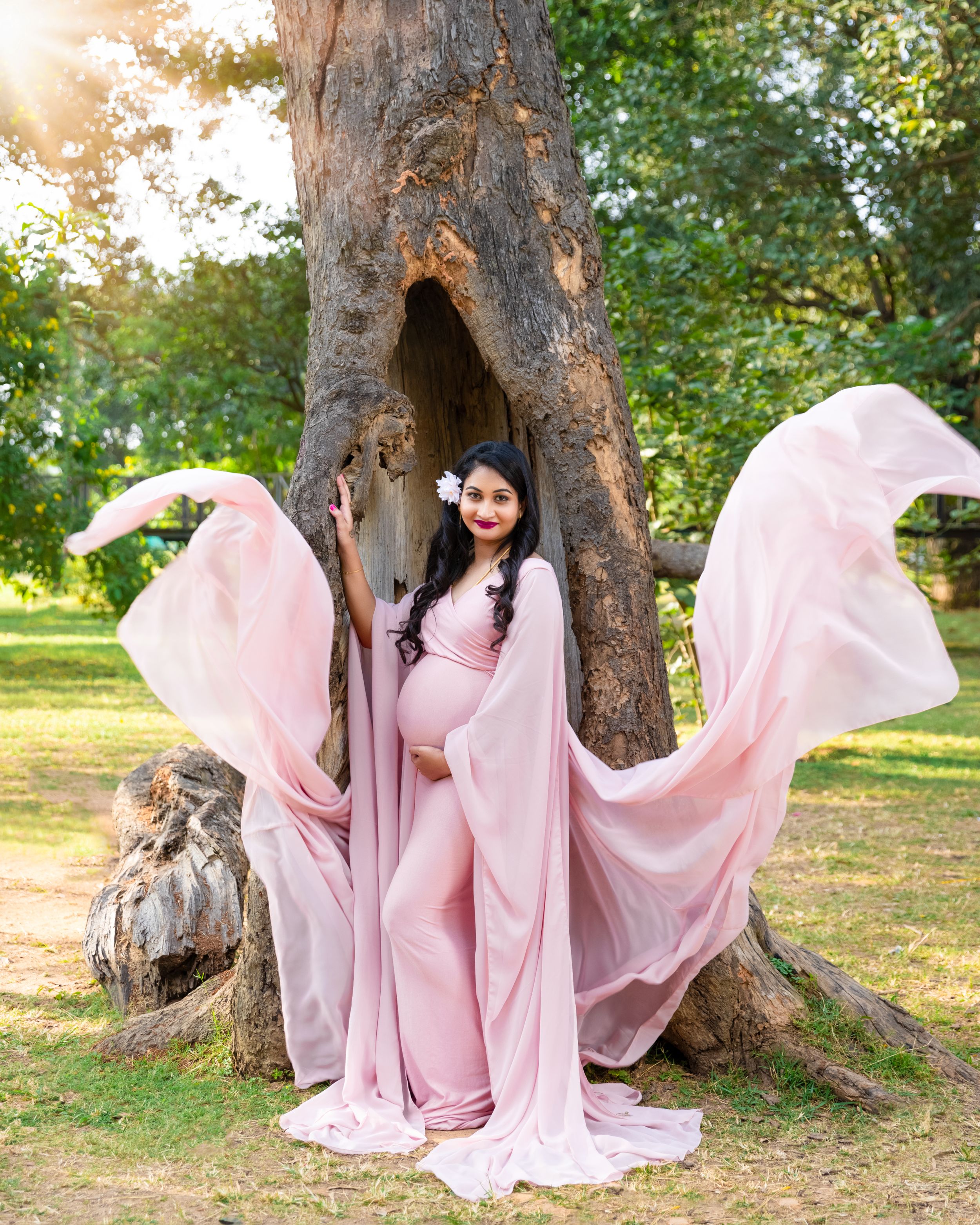 Flowing pink maternity dress photographed against a hollow tree trunk with dramatic fabric movement in golden sunlight.