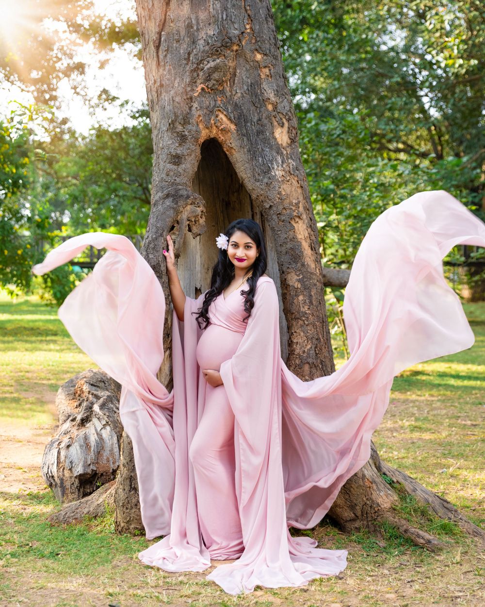Flowing pink maternity dress photographed against a hollow tree trunk with dramatic fabric movement in golden sunlight.
