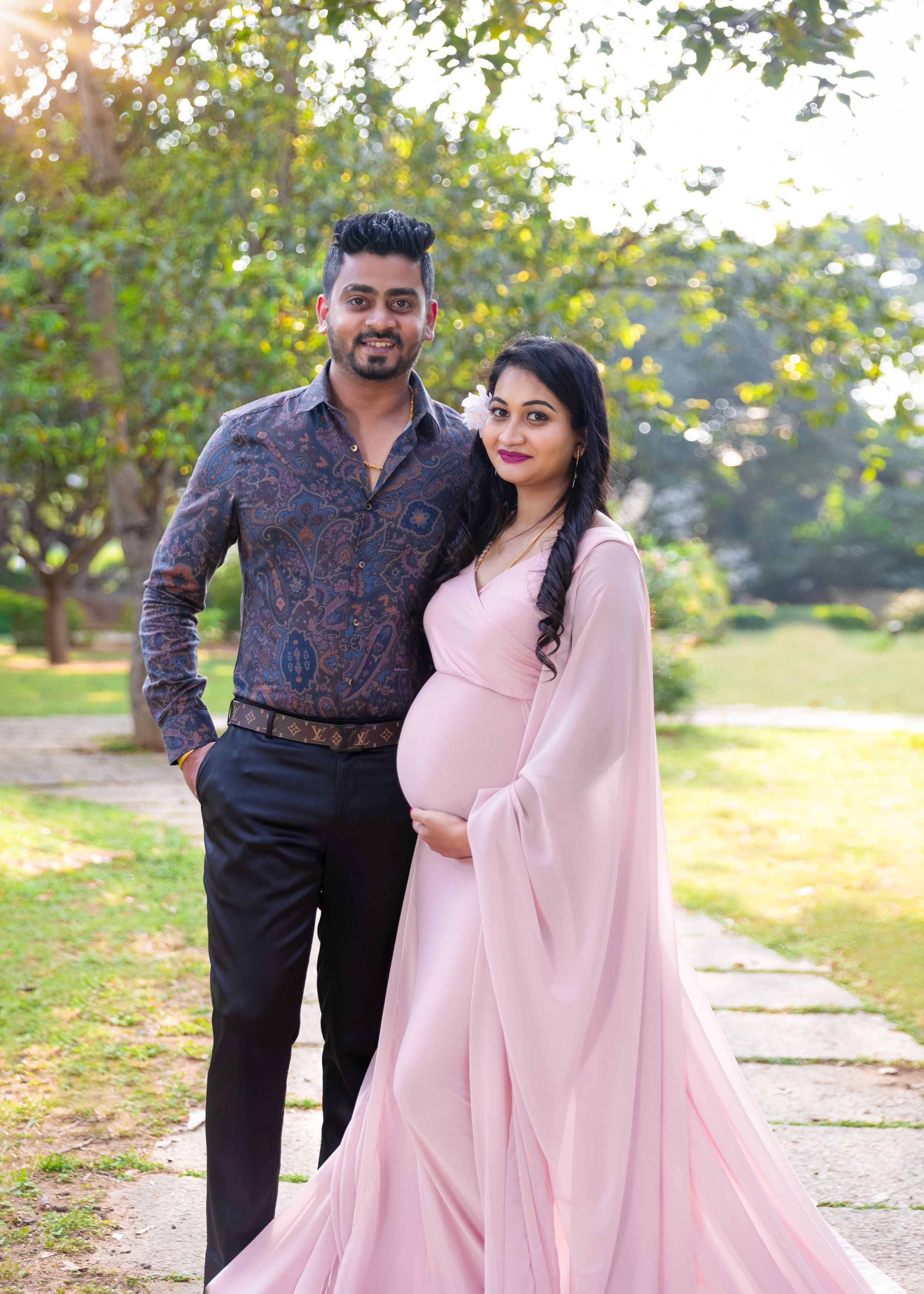 Expectant couple poses outdoors in complementary outfits with pink flowing gown and patterned shirt.