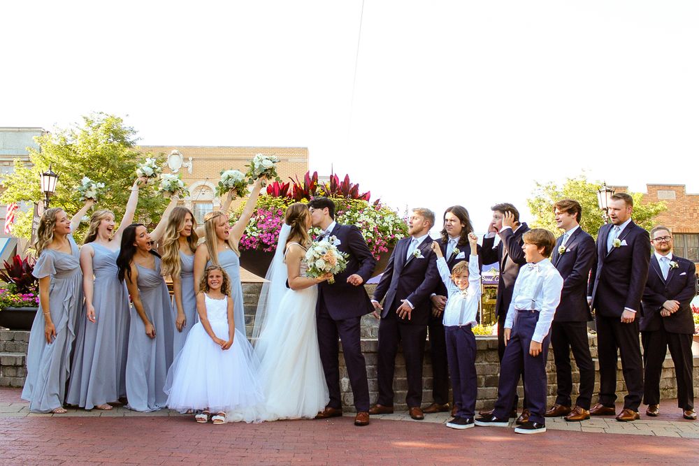 Large wedding party poses outdoors in formal attire with bridesmaids in gray dresses.