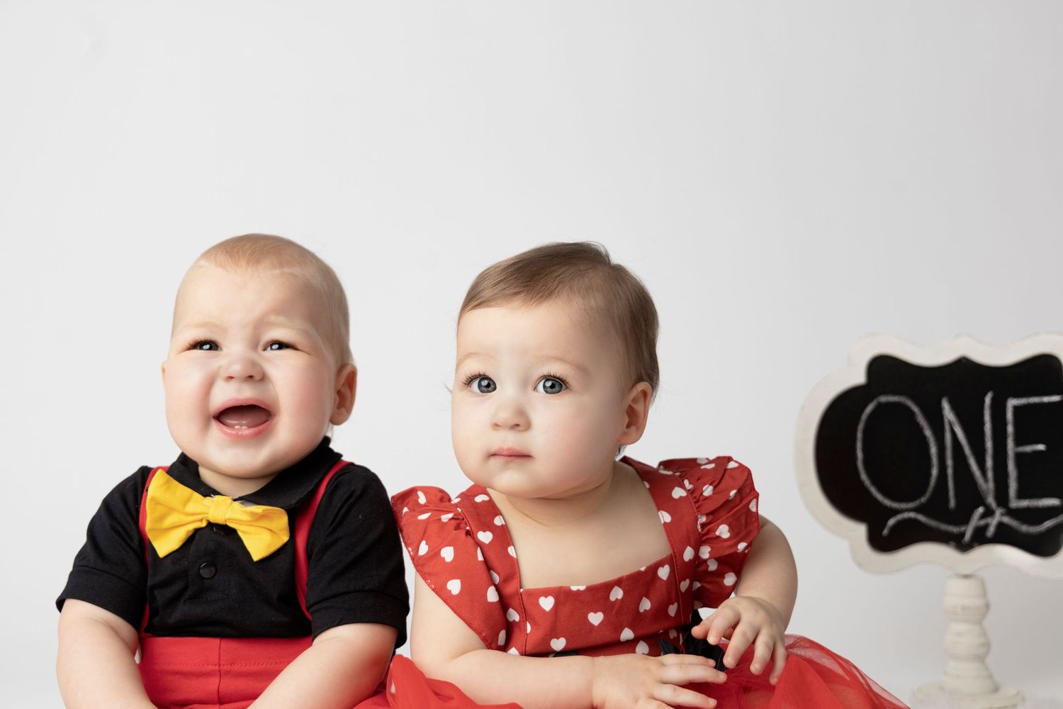 Two infants dressed in formal attire with polka dots and bowties pose together for a first birthday photo shoot.