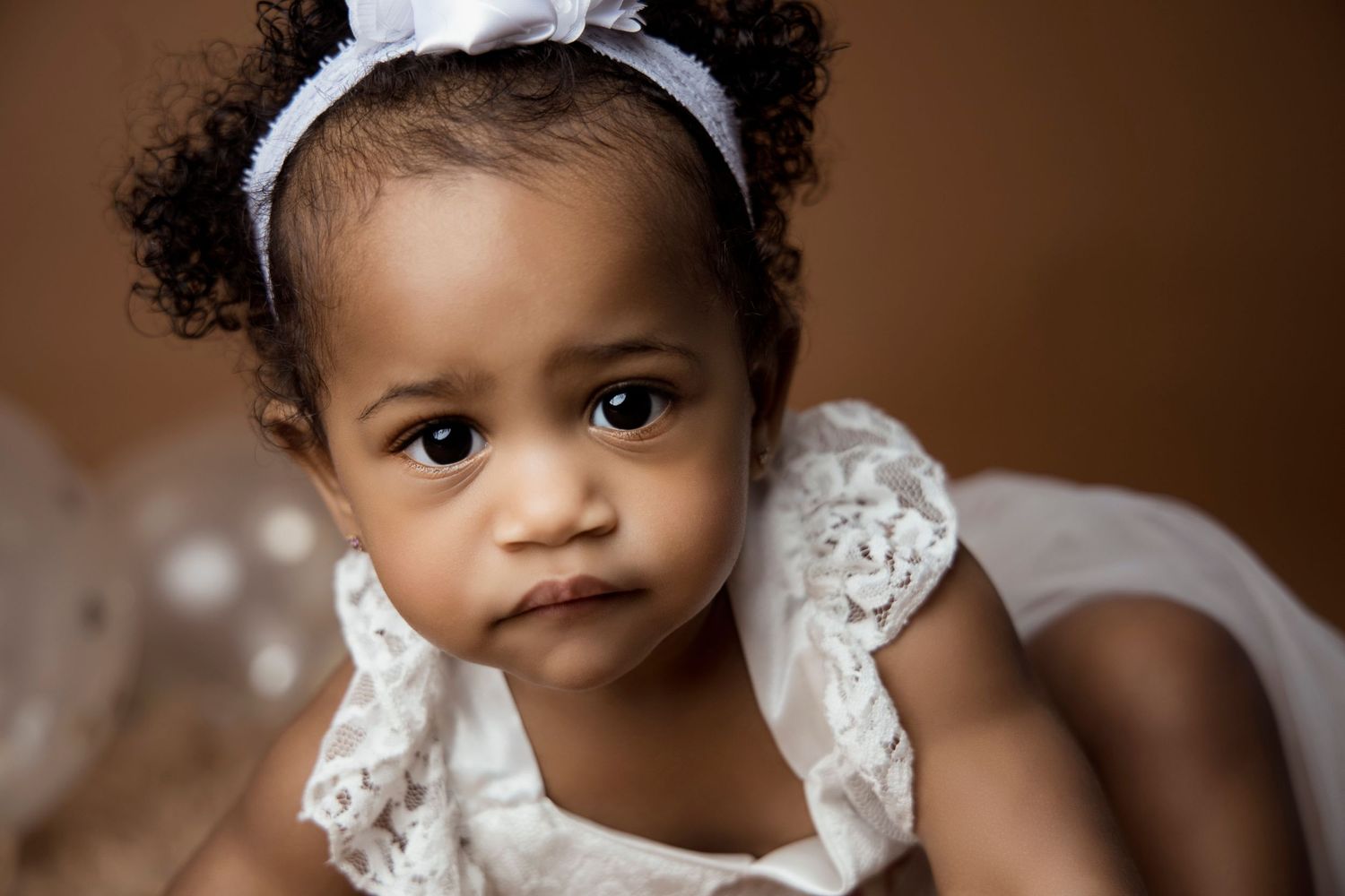 Baby in white lace dress with headband poses for portrait against brown background.