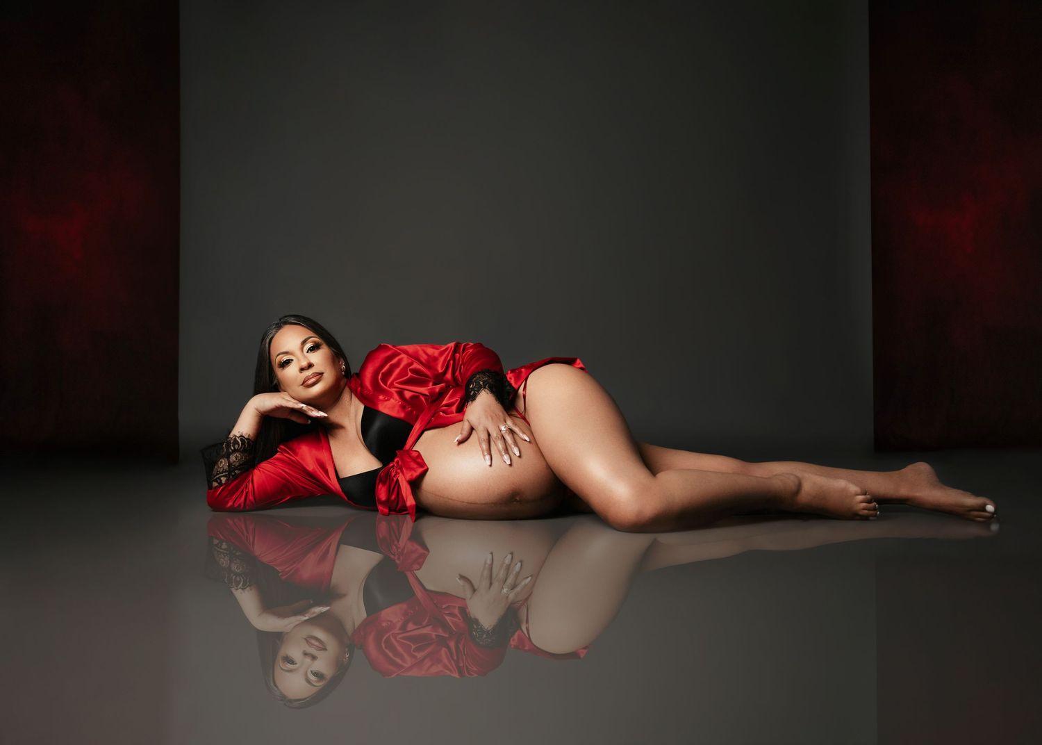 Elegant maternity boudoir photo with red satin lingerie against dark background with reflection.