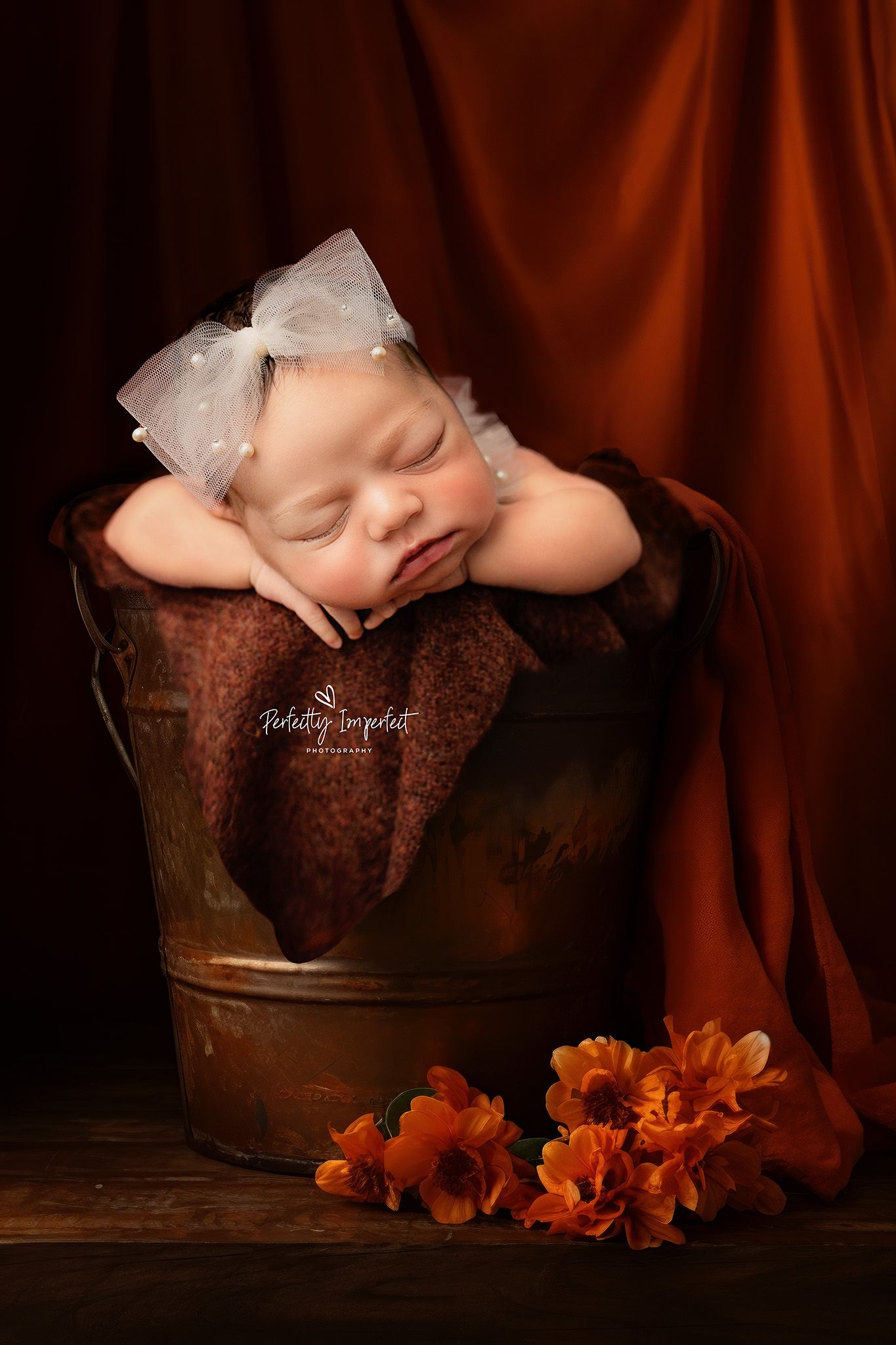 A sleeping newborn in a vintage metal bucket with orange flowers beside it against a dark brown backdrop.