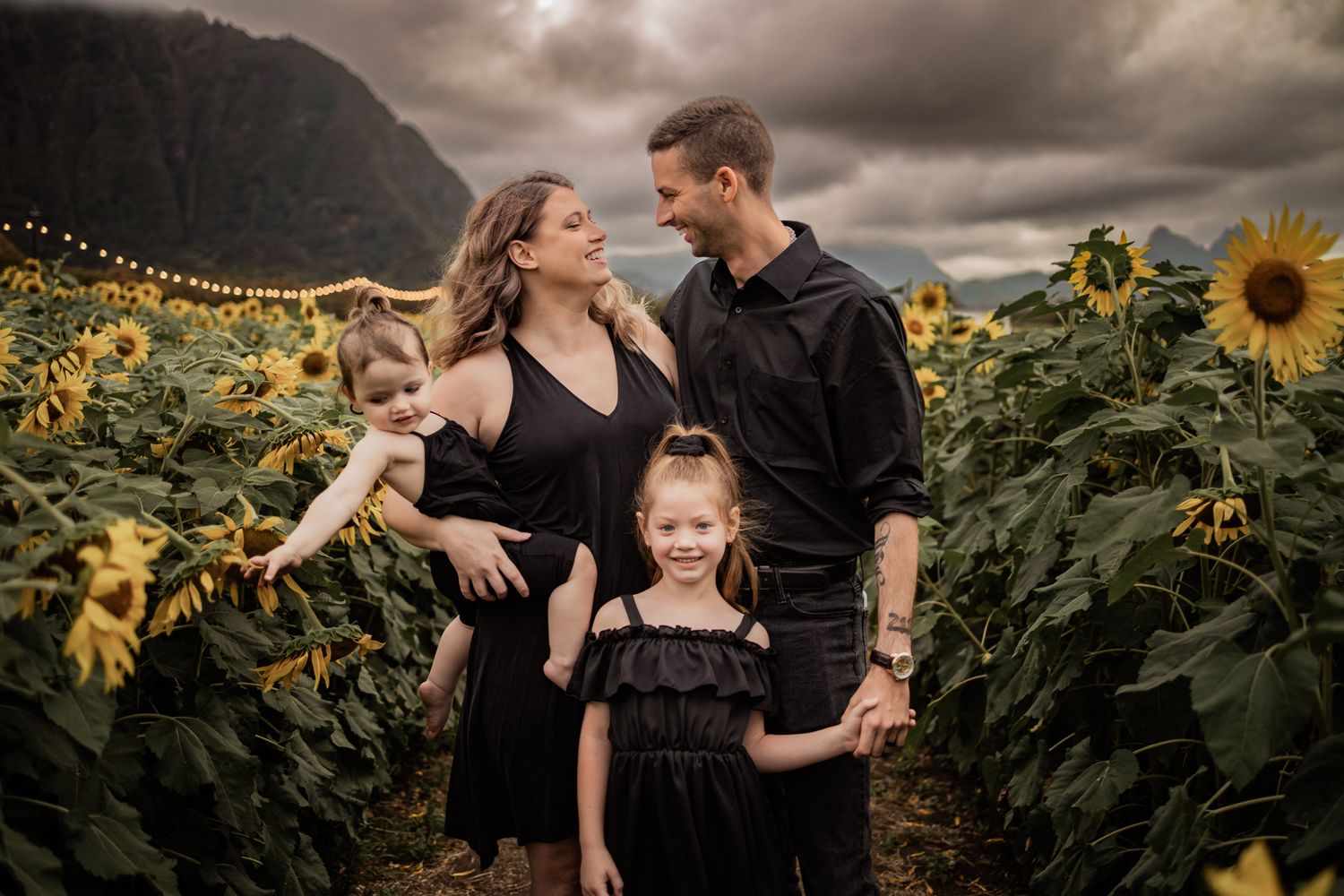 Family embraces in a sunflower field at sunset with dramatic mountain backdrop.