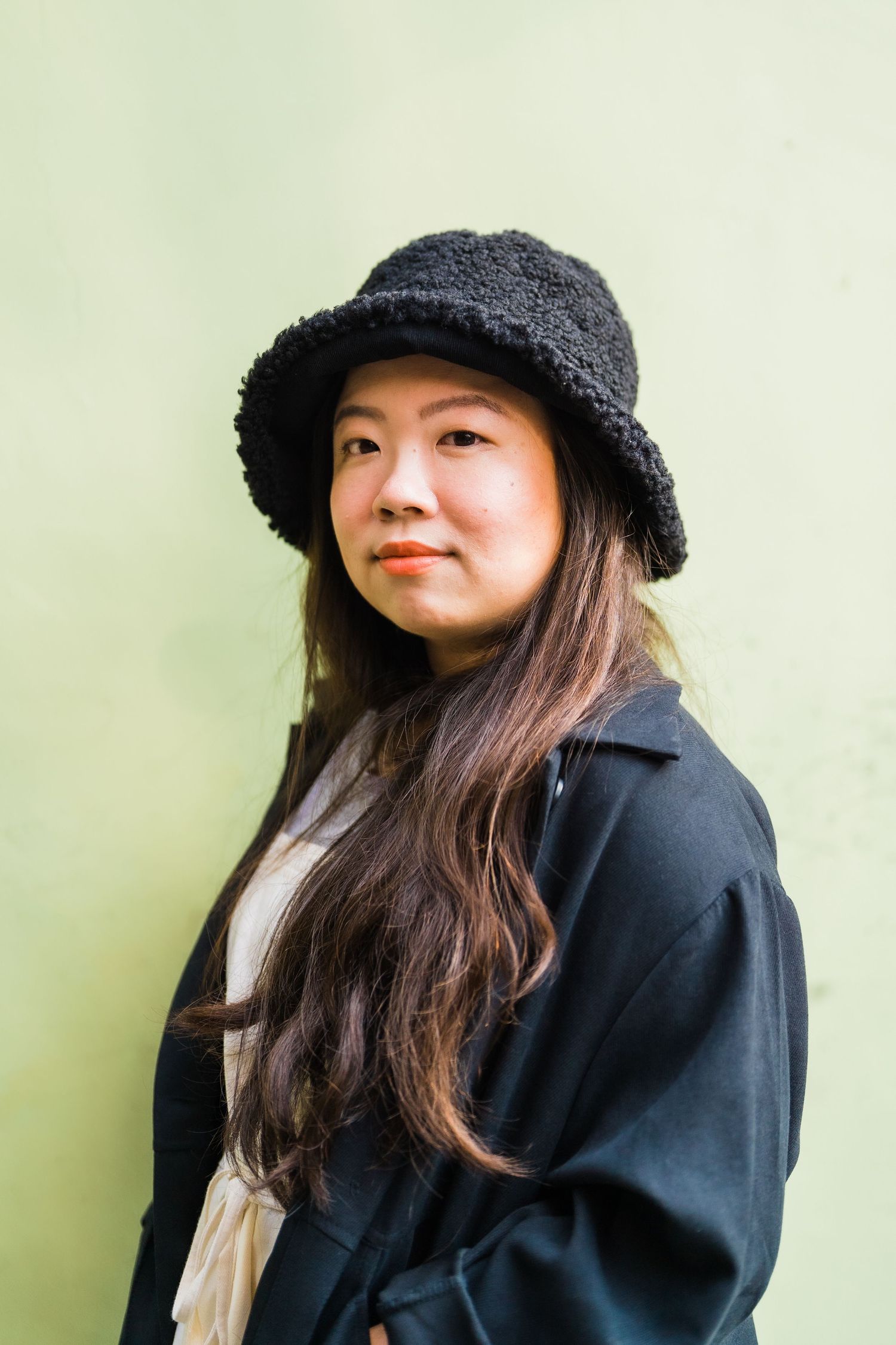 Person in black coat and fuzzy bucket hat poses against light green wall.