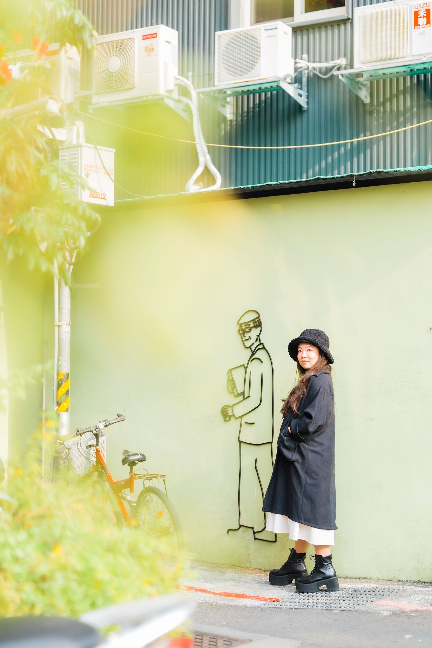 Person in black coat poses next to painted wall figure in dreamy sunlit setting.