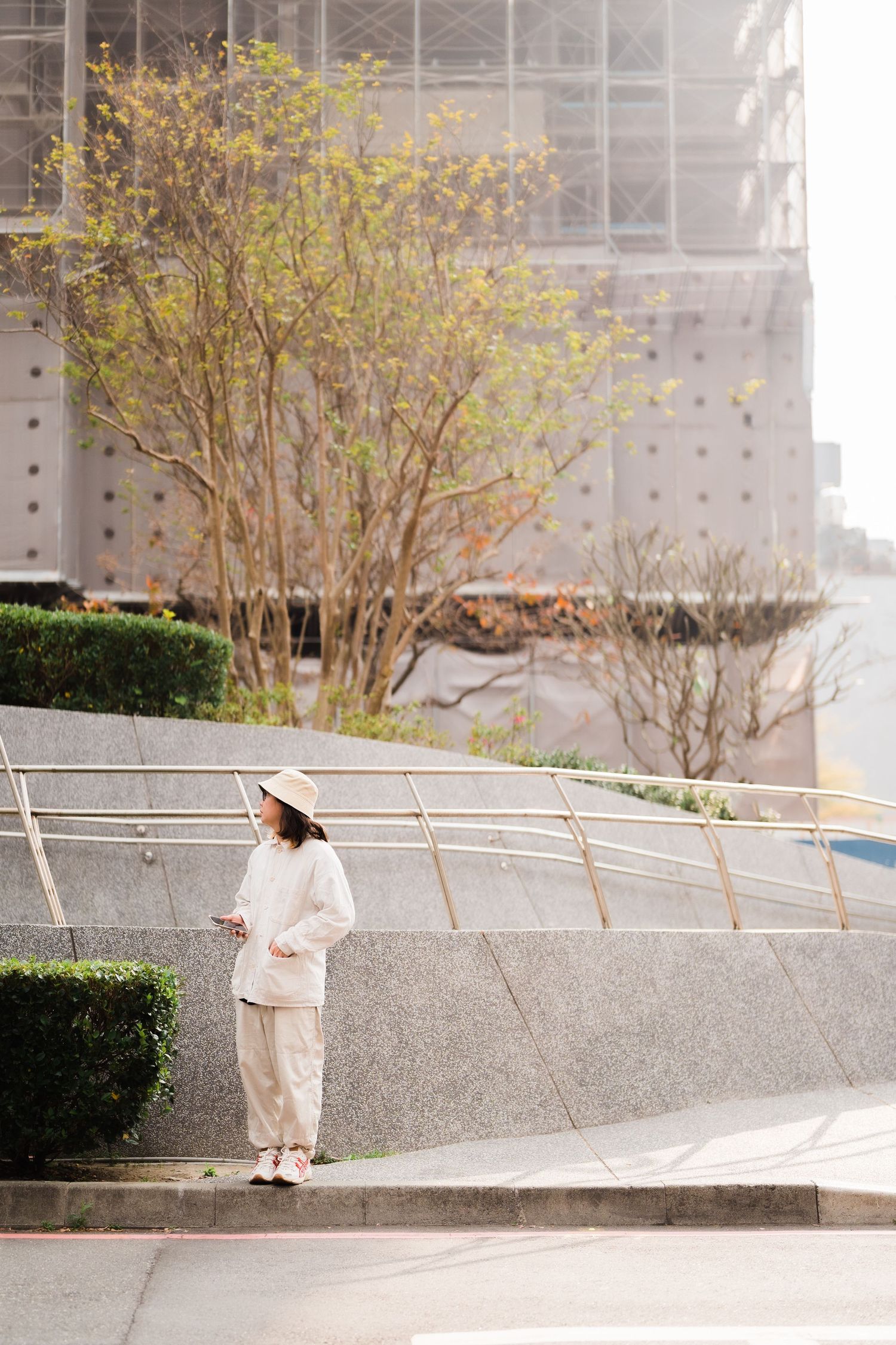 A person in white clothing stands near a yellow-leafed tree against an urban concrete backdrop.
