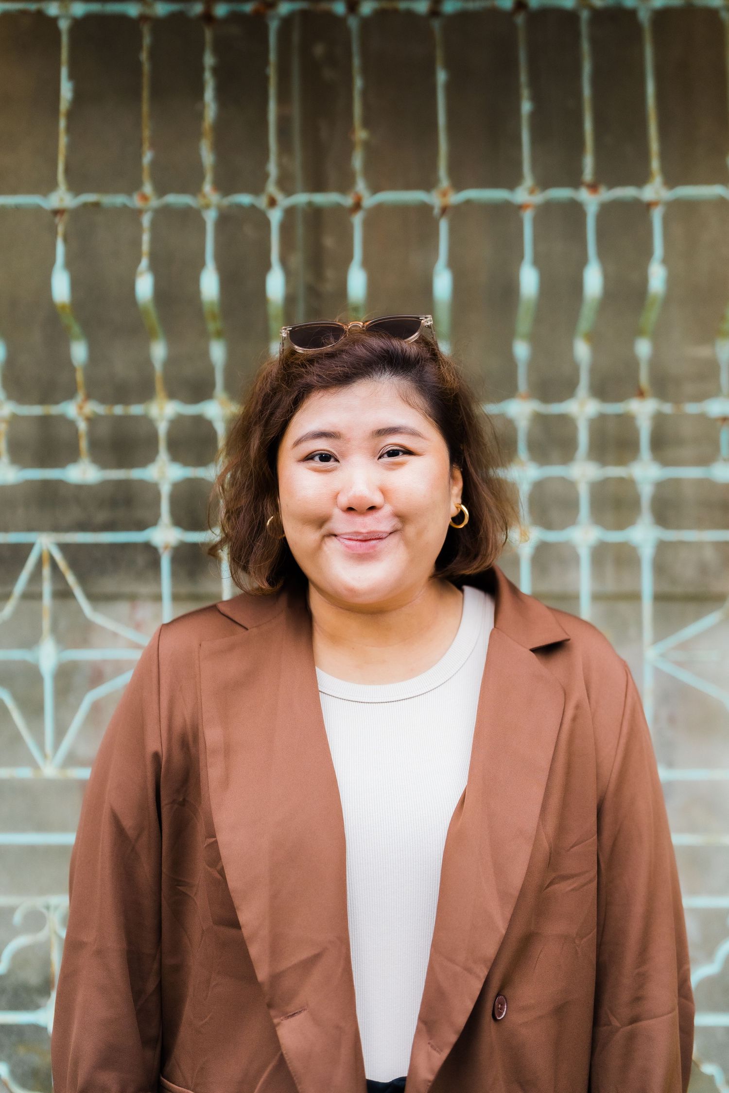 Person in a brown blazer smiling warmly against a patterned wall background.