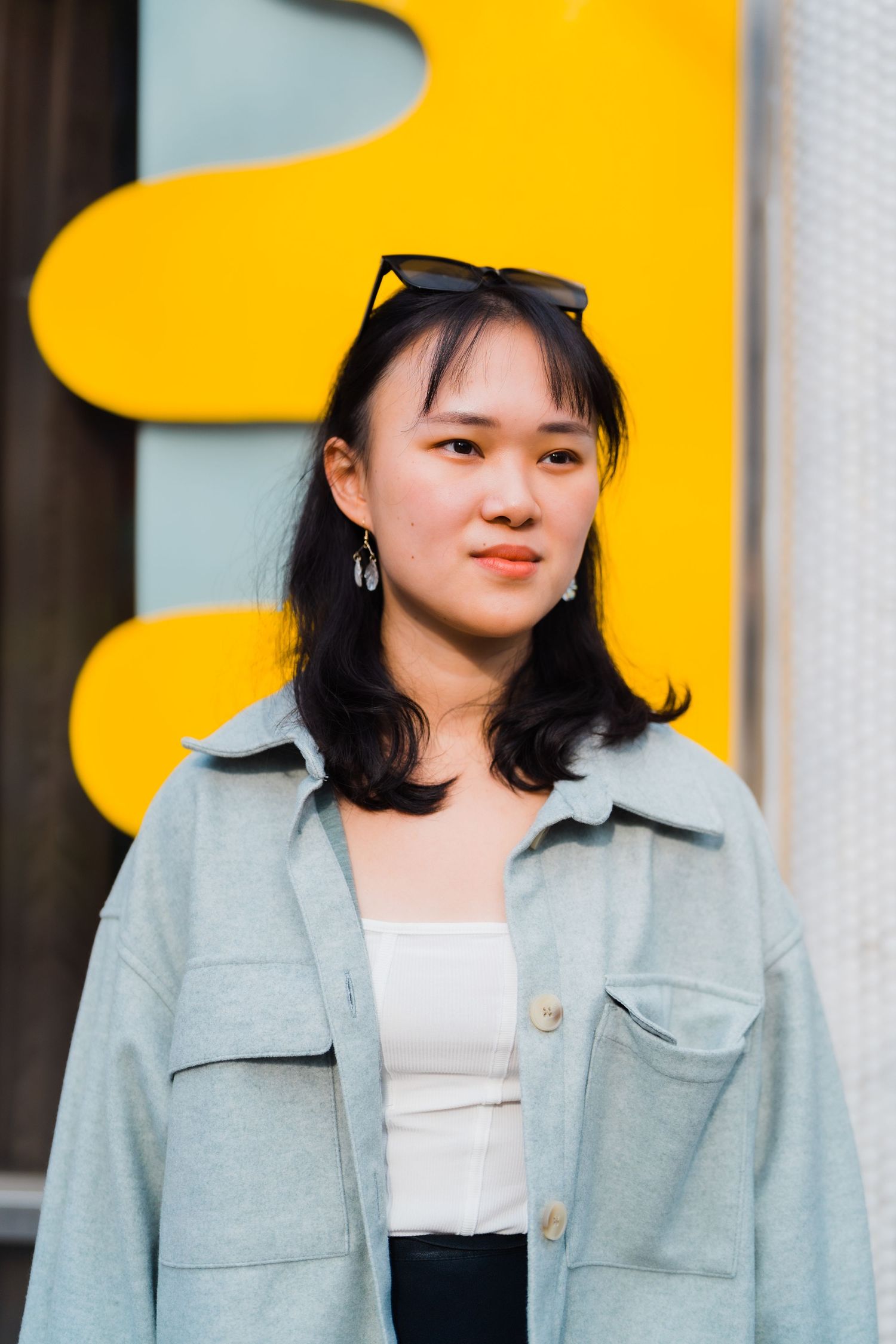 Portrait against yellow wall showing fashionable winter outfit with light blue jacket and sunglasses on head.