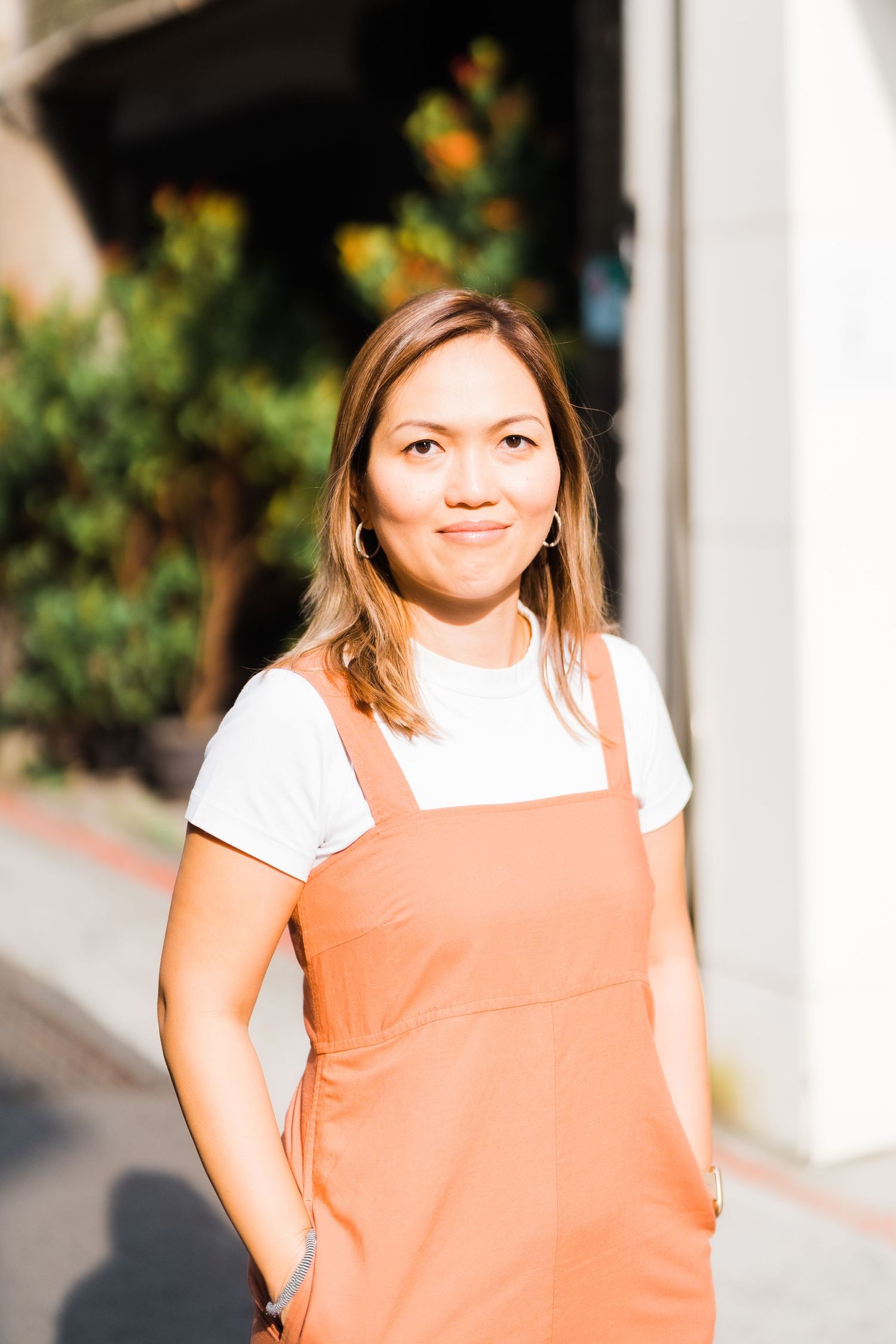 Person wearing a white t-shirt and peach overall dress stands in sunlight.