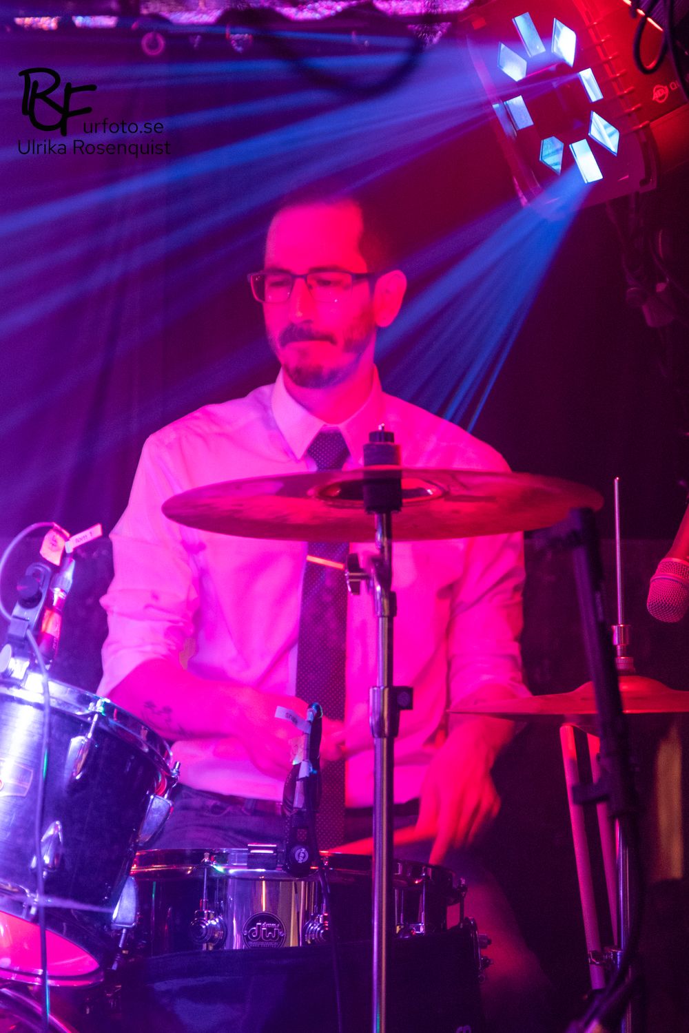 A drummer performs under vibrant purple and red stage lighting at a concert venue.