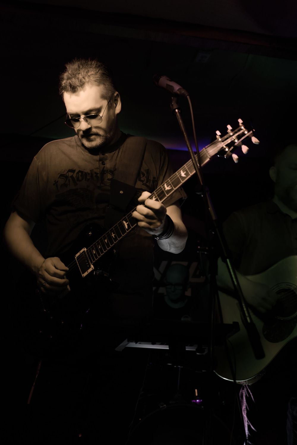 Guitarist performs on darkly lit stage, playing electric guitar in dramatic lighting.