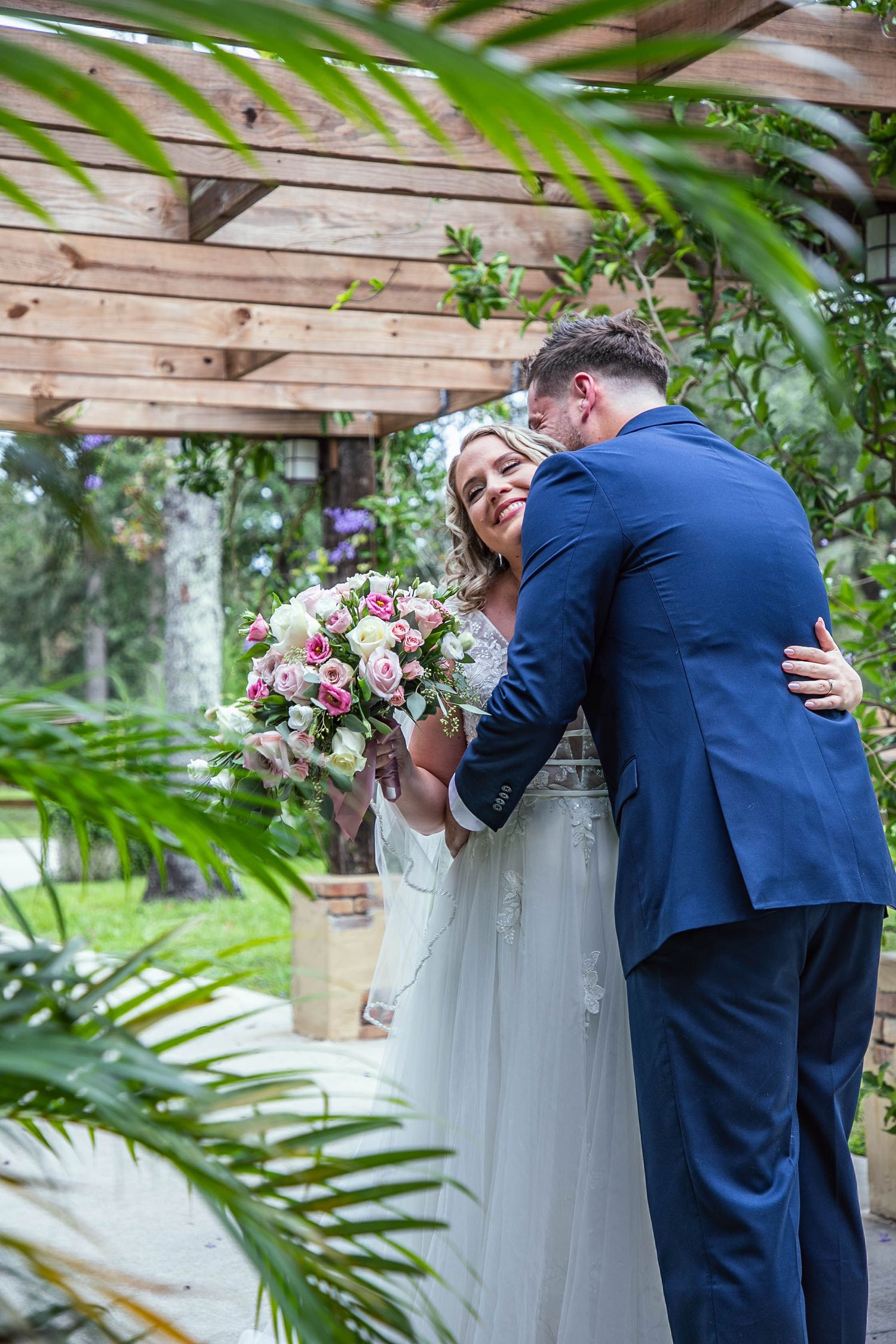 Couple embracing under wooden pergola with pink rose bouquet and tropical foliage.