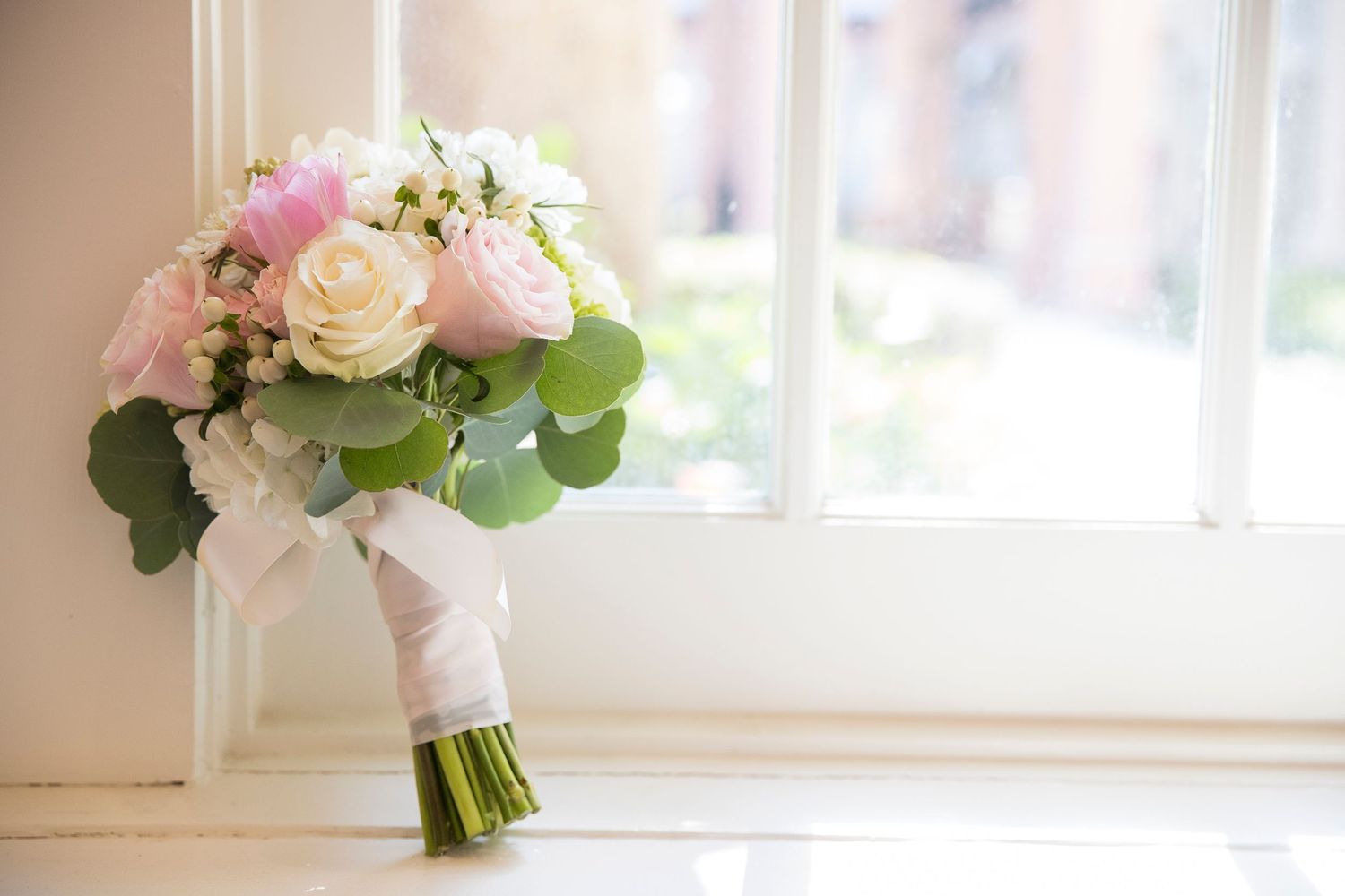 Elegant bridal bouquet with pink and white roses and eucalyptus leaves by window.