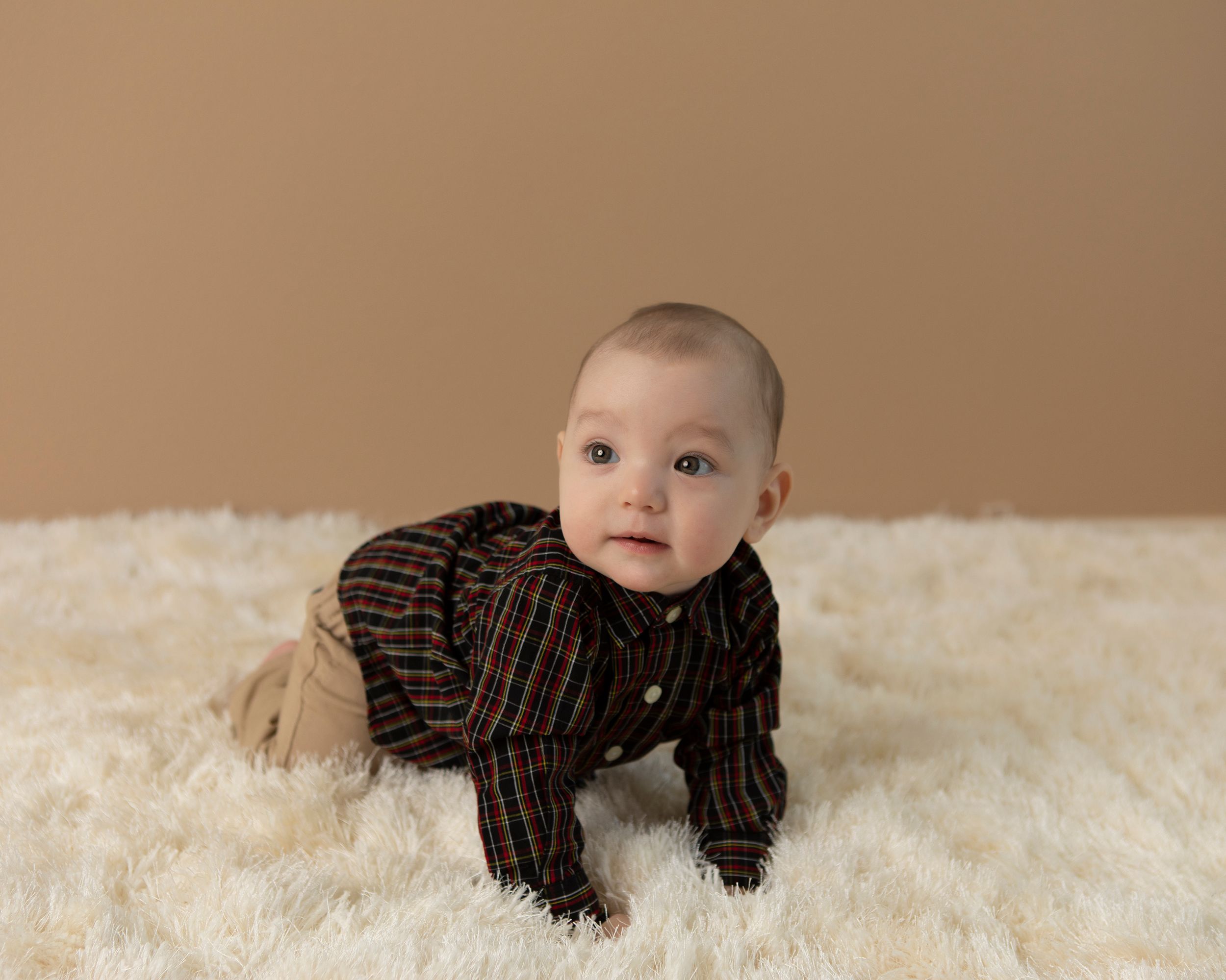 Baby in plaid outfit crawling on white furry carpet during professional photo session.