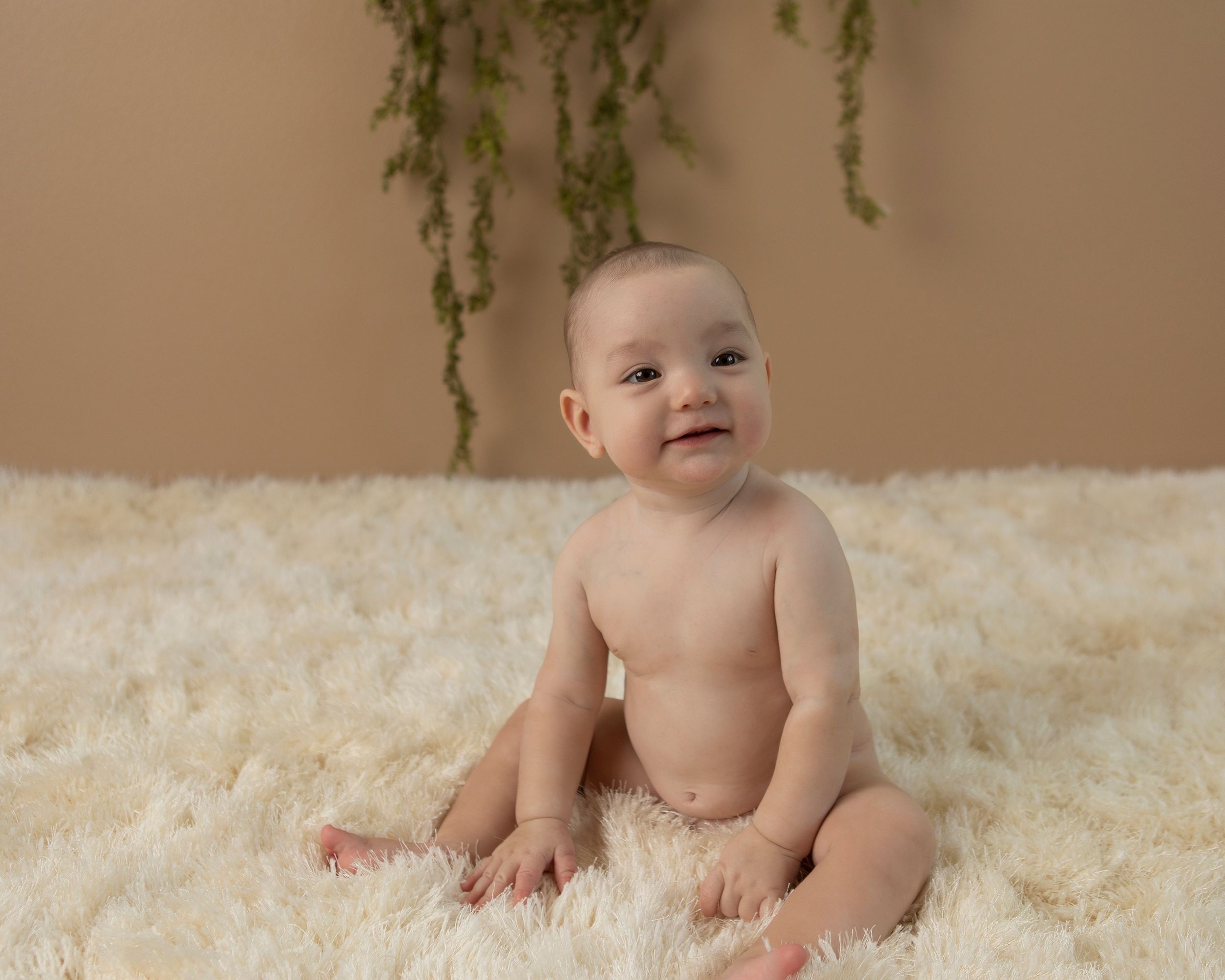 Baby sitting on white fluffy rug with hanging greenery in background during photo session.