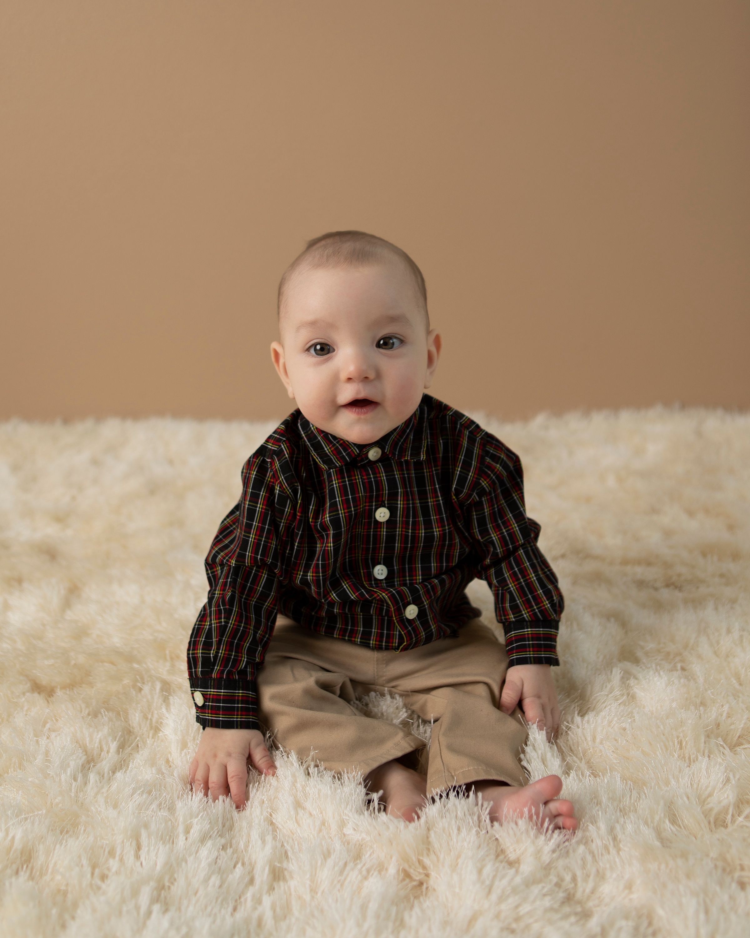 Baby in striped dress shirt and khakis sitting on white furry rug against tan background.