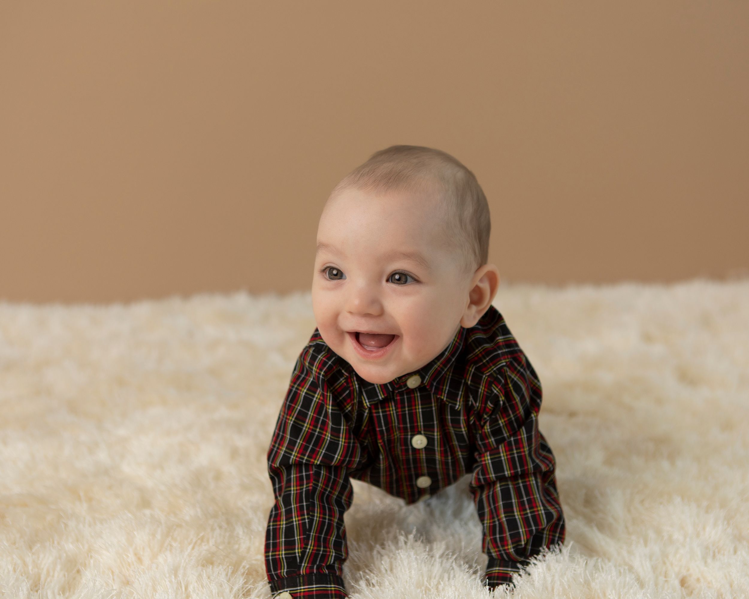 Baby wearing plaid button-up shirt smiling while lying on white fluffy rug against brown background.
