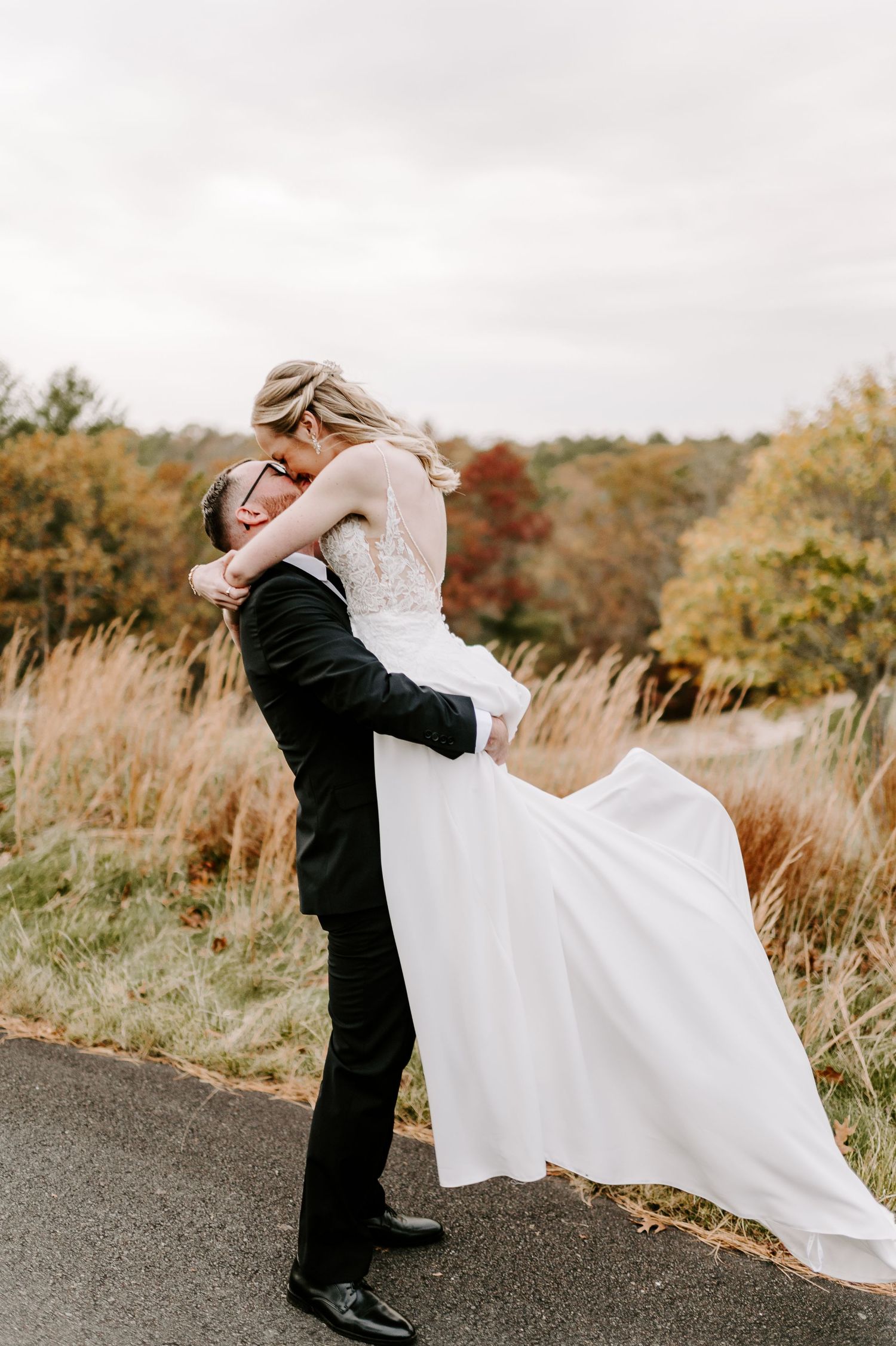 A groom lifts his bride in a flowing white dress on a winding path amid fall foliage.