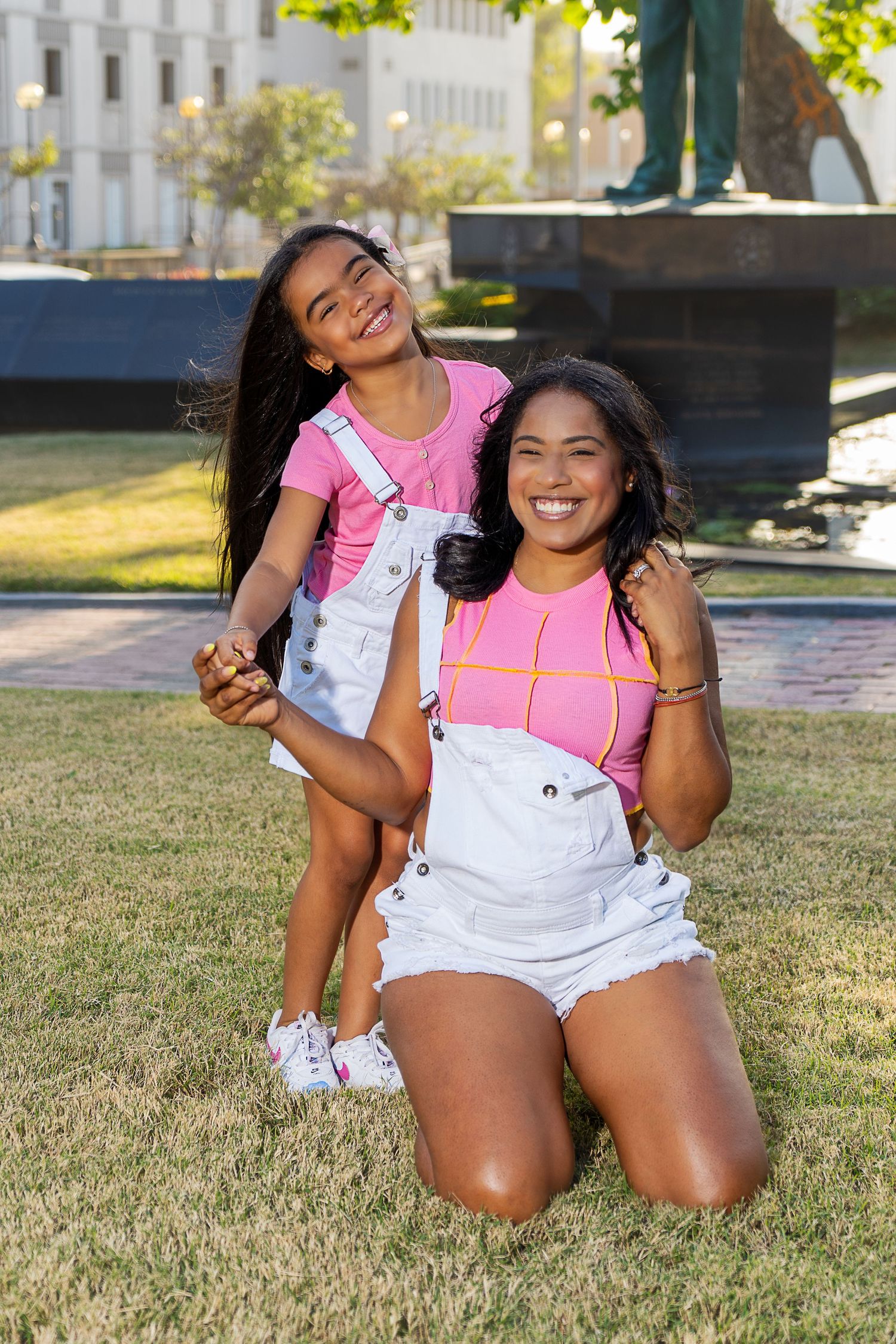 Playful portrait in matching pink tops and white overalls on sunny grass lawn.