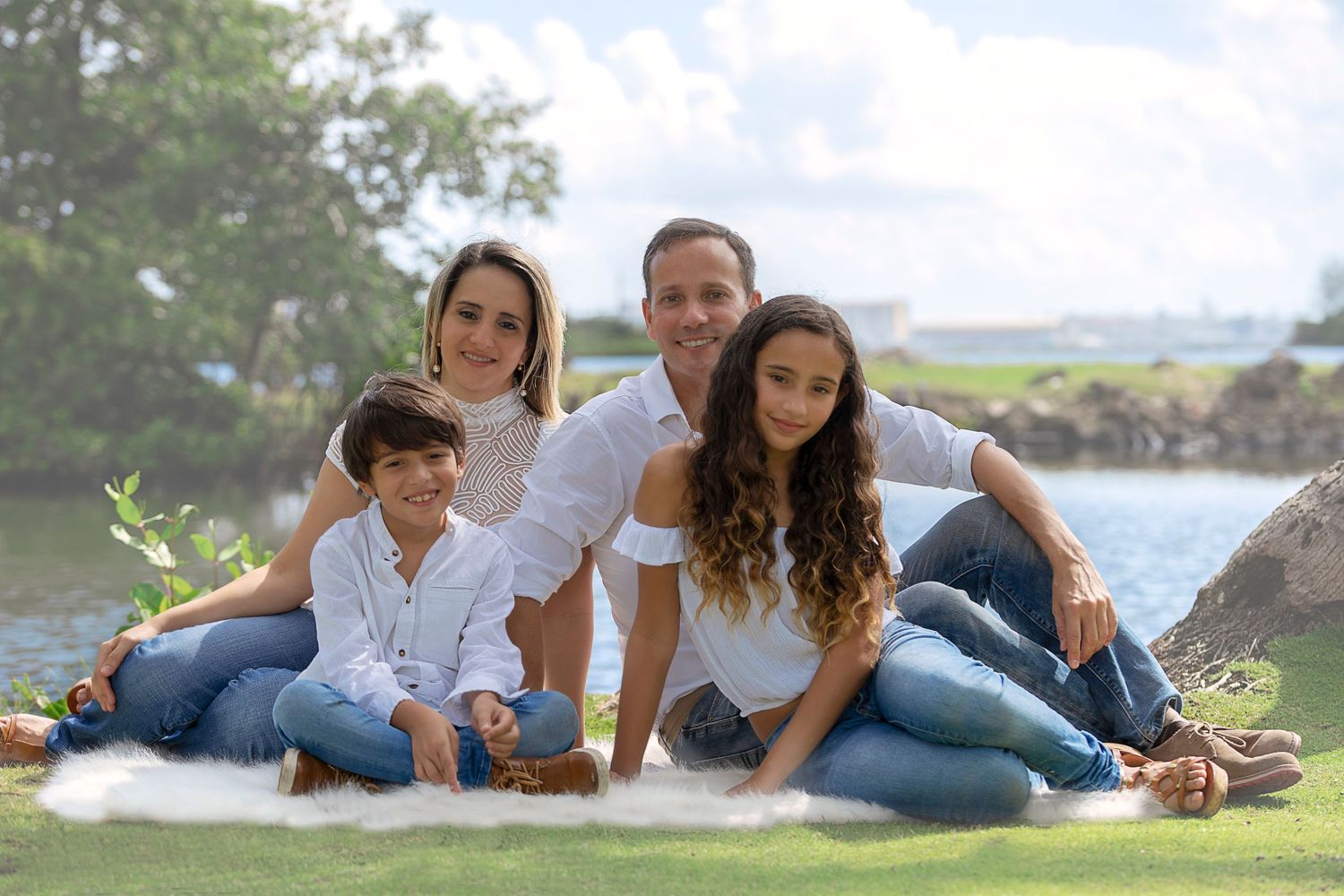 A family in coordinated white and denim outfits relaxes together on a grassy hillside.