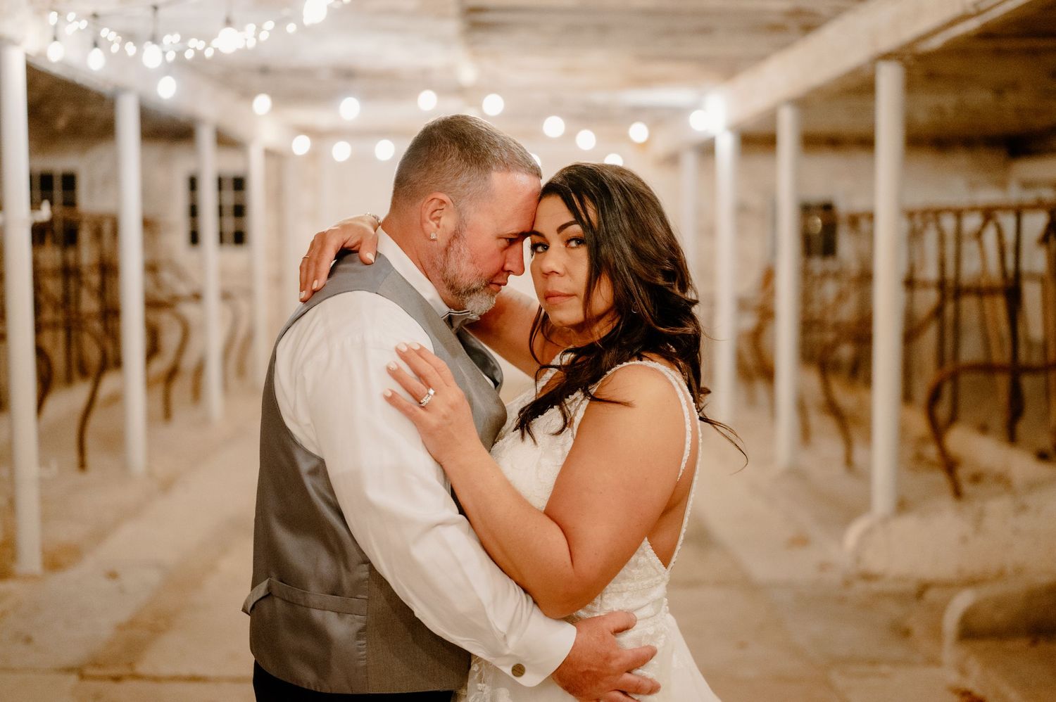 An intimate moment between newlyweds in a rustic venue with string lights overhead.
