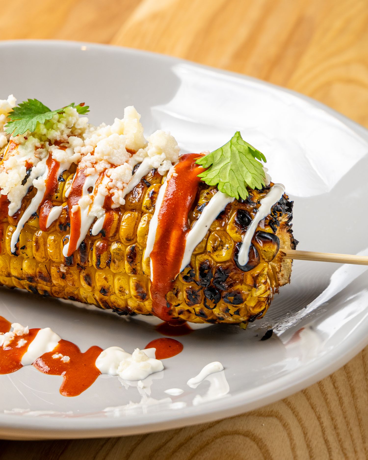Grilled corn on the cob topped with drizzles of white sauce and red sauce, garnished with fresh cilantro on a white plate.