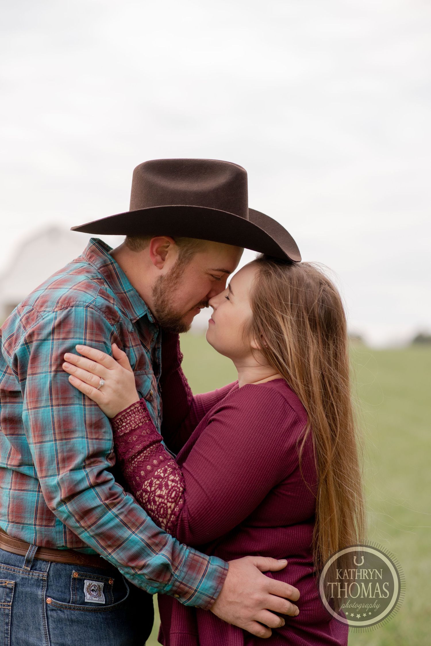 A couple shares an intimate moment in a field wearing western attire with a brown cowboy hat.