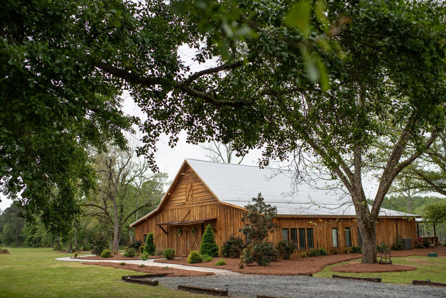 Rustic wooden barn and venue building nestled under large shade trees at a country event space.