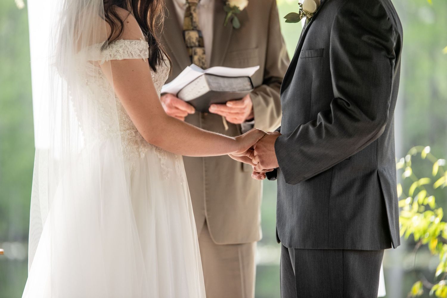 A couple holding hands during an outdoor wedding ceremony as they exchange vows with an officiant holding a bible.