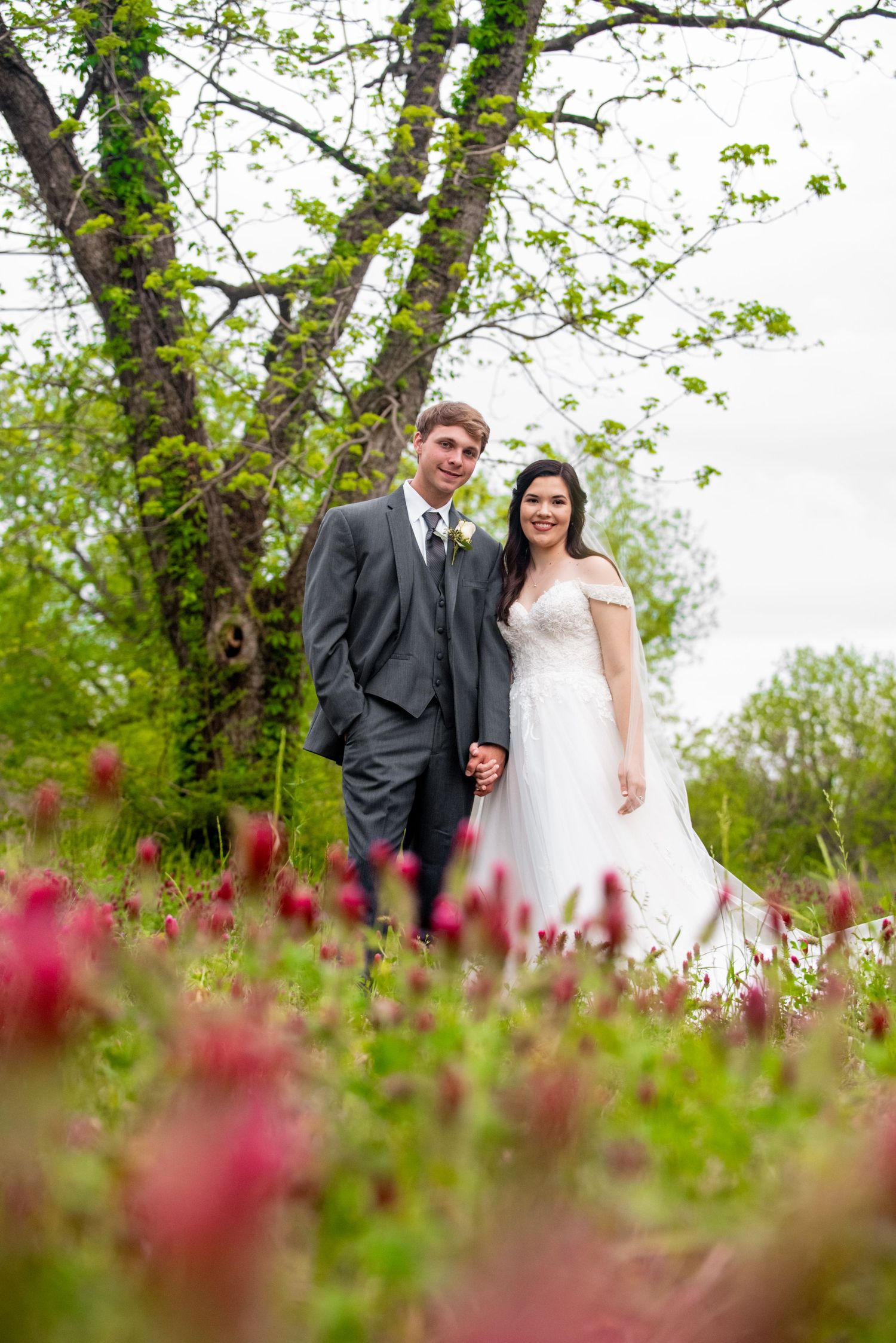 A couple stands amid spring foliage and pink flowers on their wedding day in a natural outdoor setting.