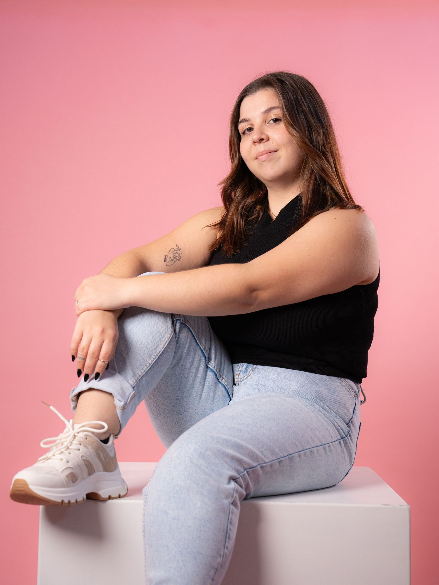 A person in casual attire sits posed on a white cube against a pink backdrop.