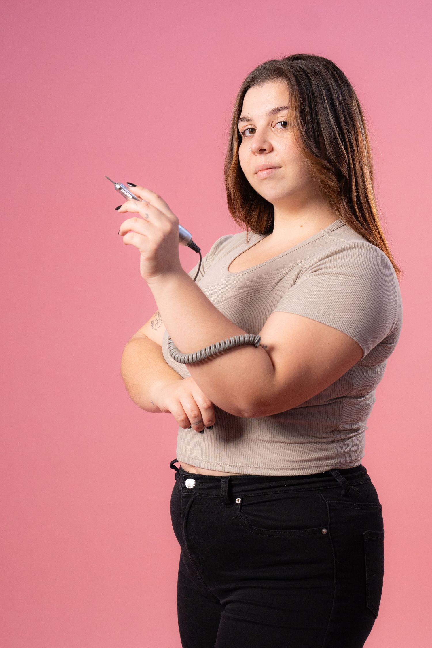 Nail technician poses confidently with tools against pink background.