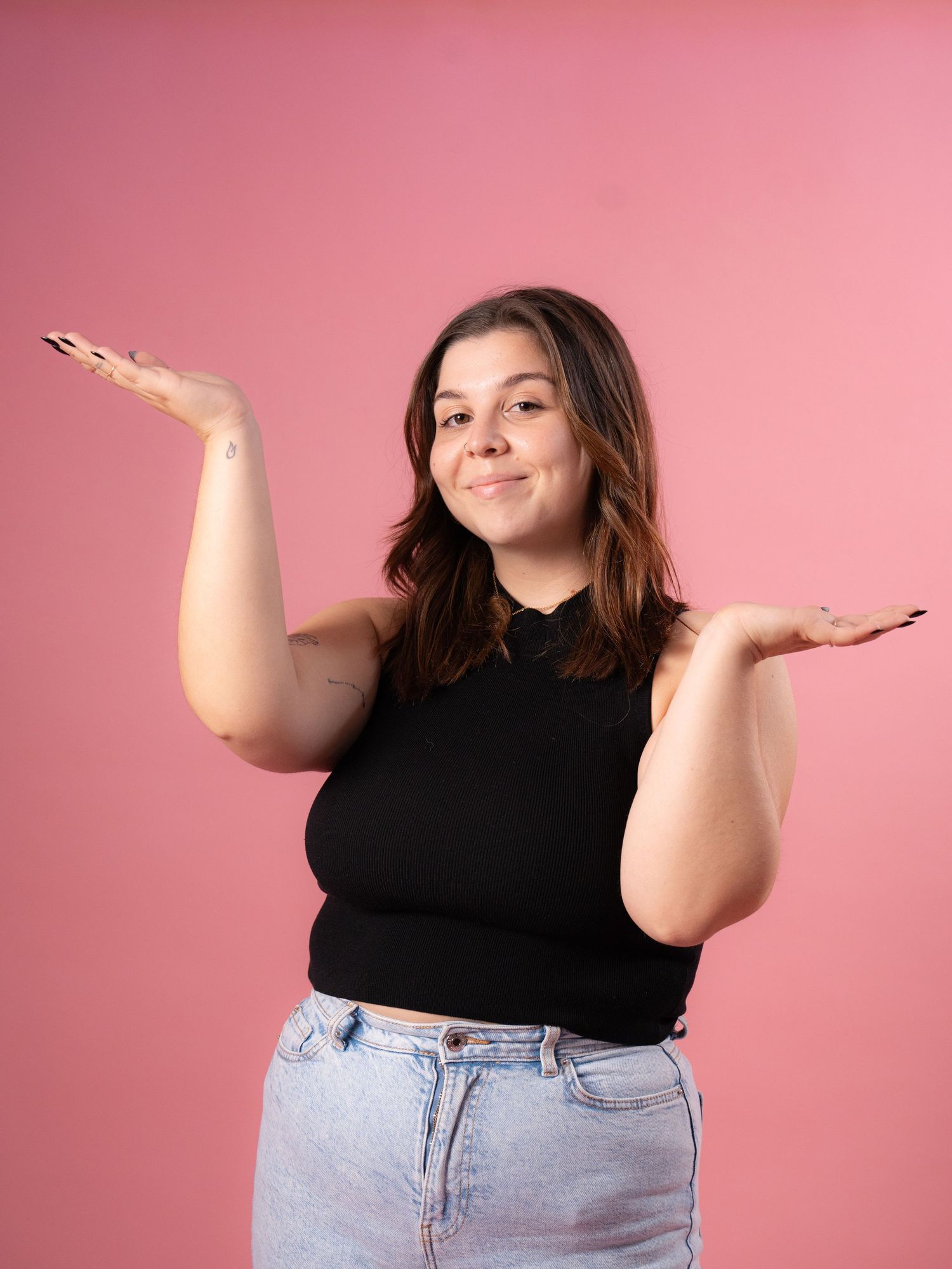 A person in black crop top and jeans gestures with raised hands against pink background.