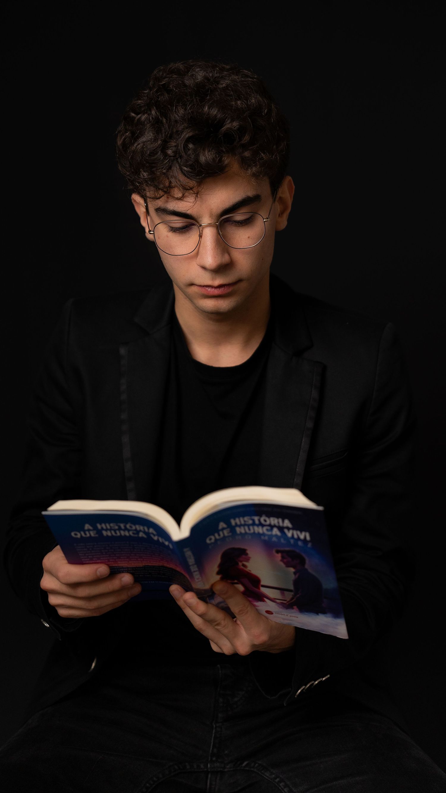 Person in dark clothing reads book intently against black background.
