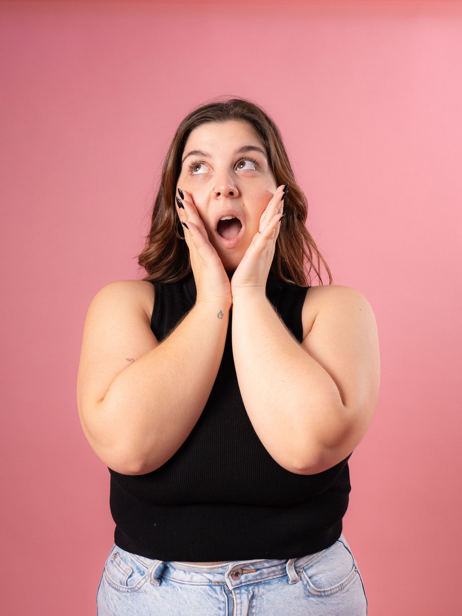 A person in a black top and jeans poses with shocked expression against a pink backdrop.
